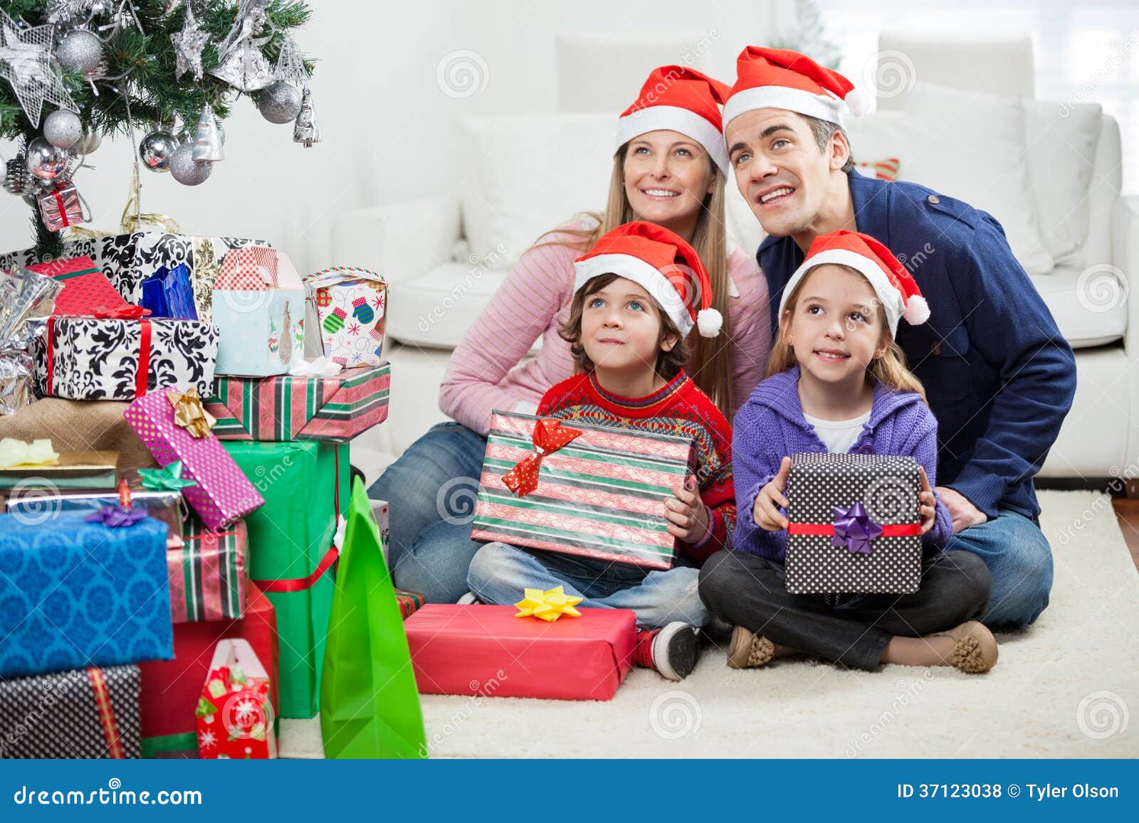 De familie in Santa Hats Sitting By Christmas stelt voor. Familie die in Kerstmanhoeden weg terwijl het zitten door Kerstmis stelt thuis voor de kijken