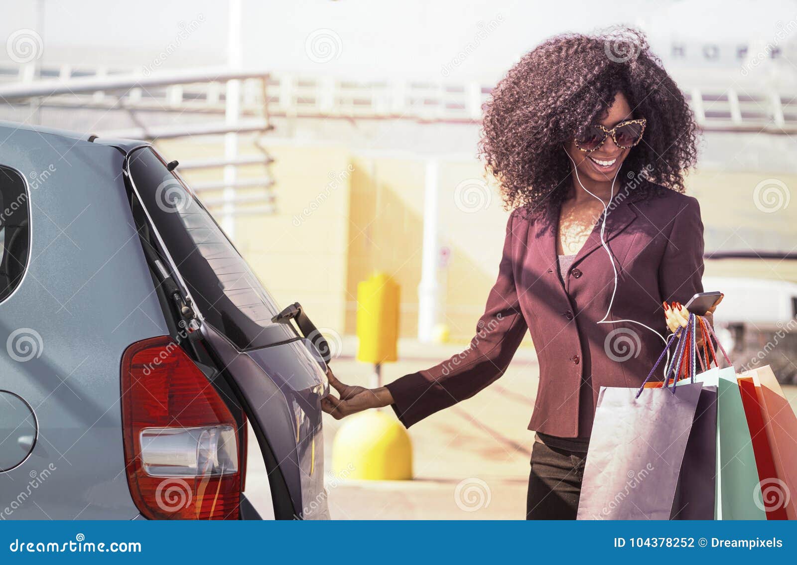 De gelukkige Afrikaanse vrouw met het winkelen doet het openen auto in zakken en het texting op mobiele telefoon. Het vrij modieuze zwarte die en haar auto met het winkelen glimlachen openen doet in hand het gebruiken smartphone in zakken
