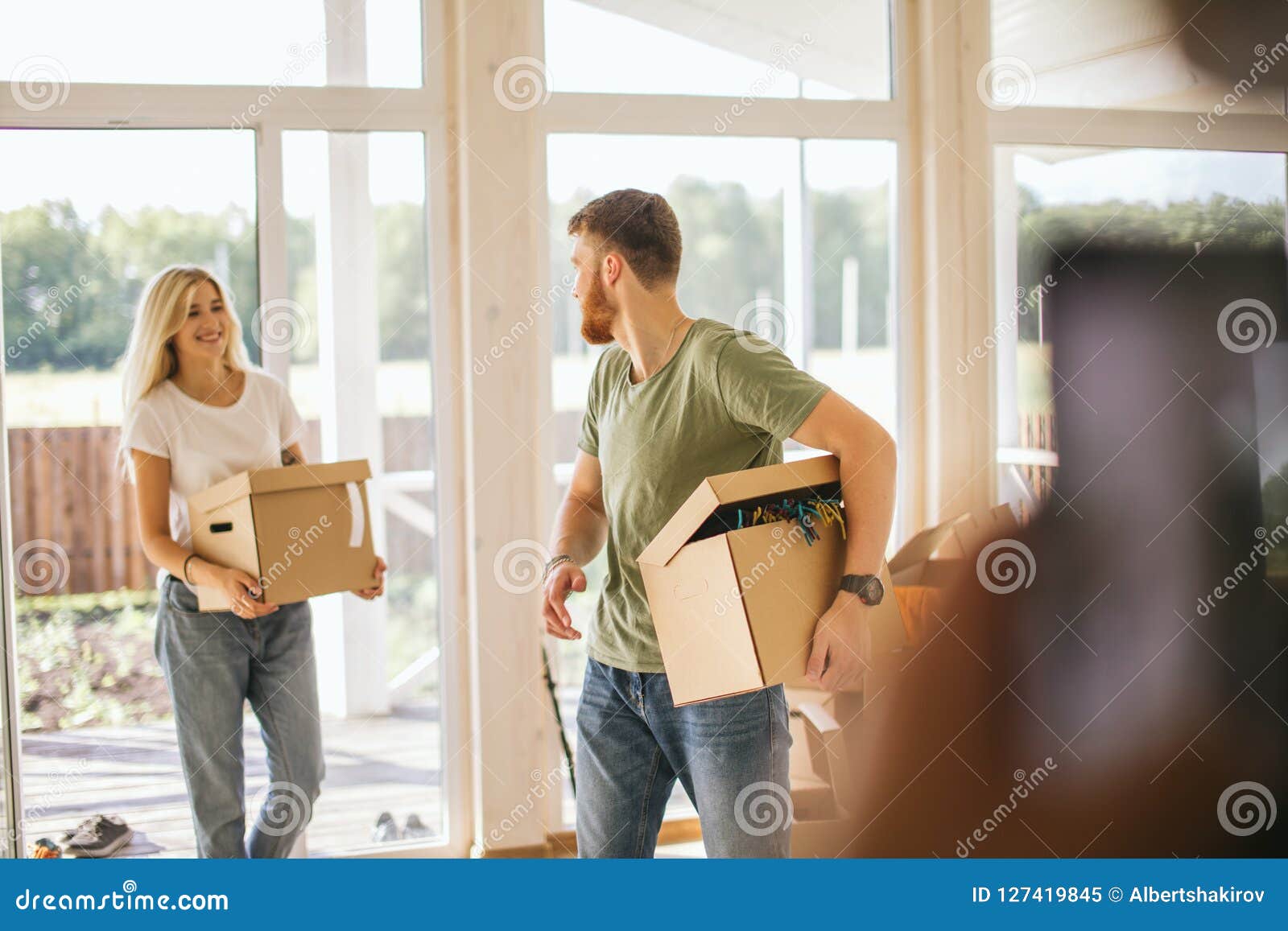 De gelukkige Dozen van het Paar Dragende Karton in Nieuw Huis bij het Bewegen van Dag. Het bewegen zich aan nieuw huis Gelukkige het kartondozen van de paarholding terwijl het gaan in het huis