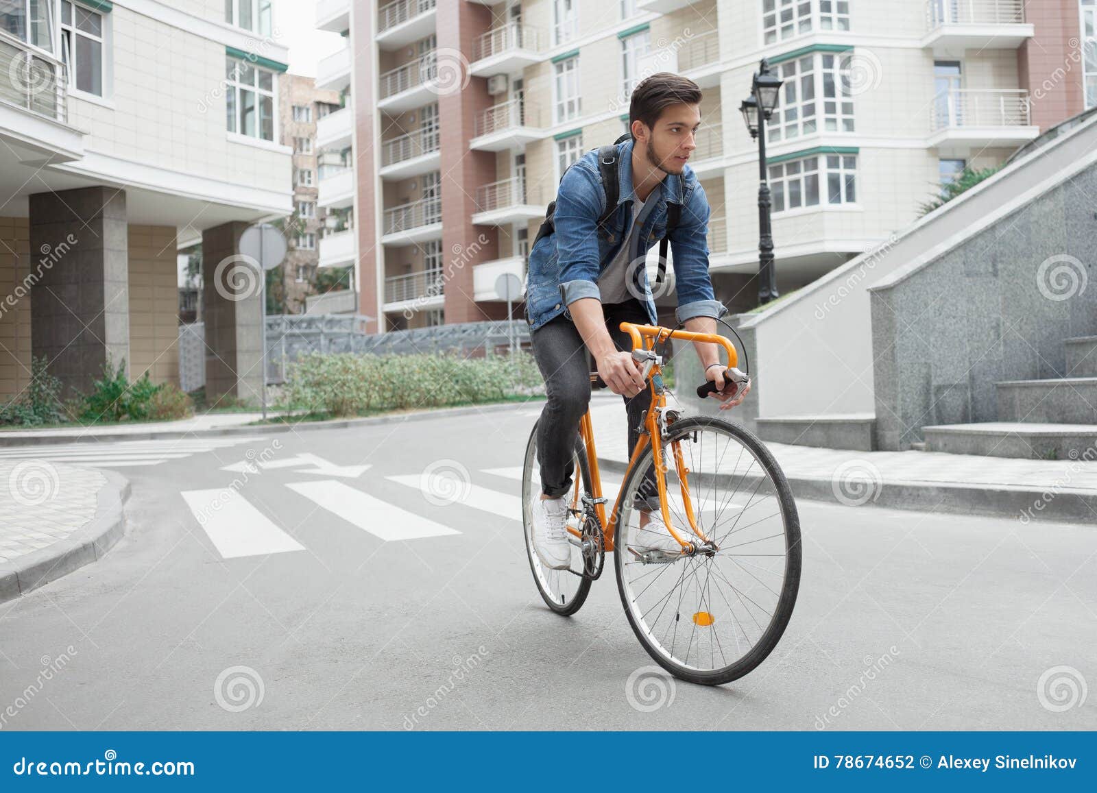 De kerel in denimjasje gaat naar school op fiets Fietsers de weg in de stad. Een kerel in een blauw denimjasje gaat naar school op een oranje fiets Fietsers op de weg in de stad Modieuze student met een rugzak