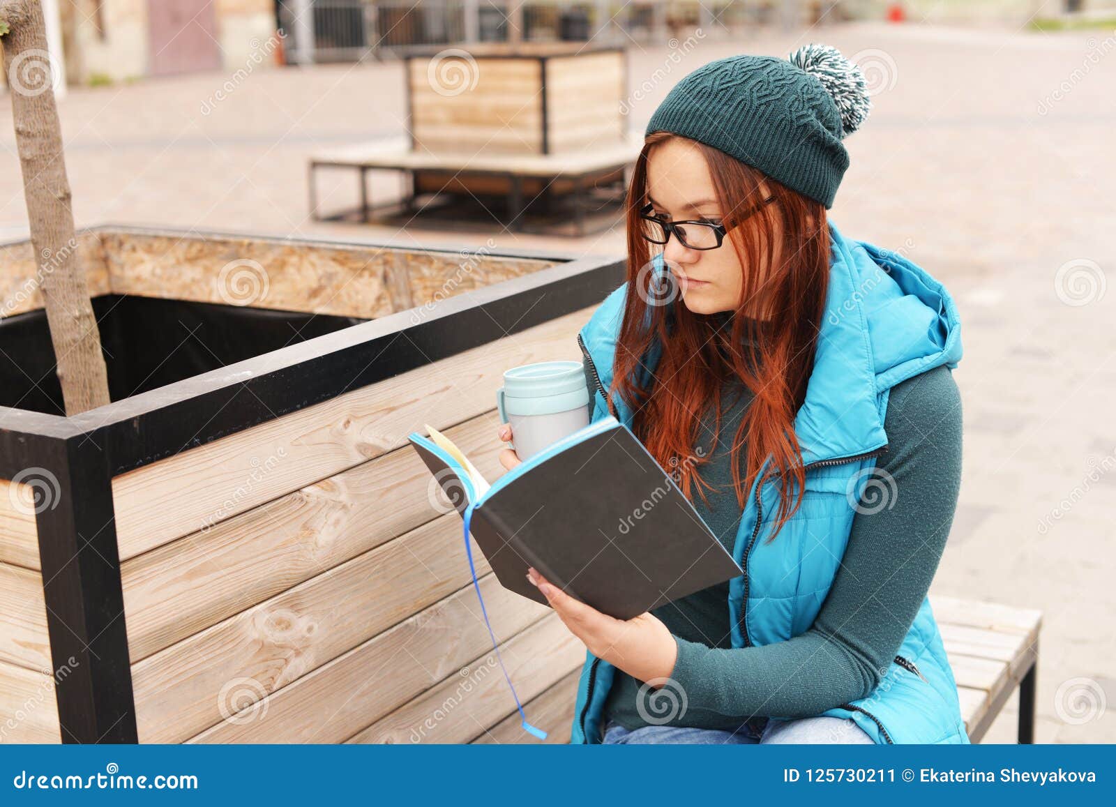 De mooie zitting van de meisjes jonge student in openlucht in een park in openlucht in de winter in de herfst en lezing een kingu in een hoed met een kop van koffie of thee in handen