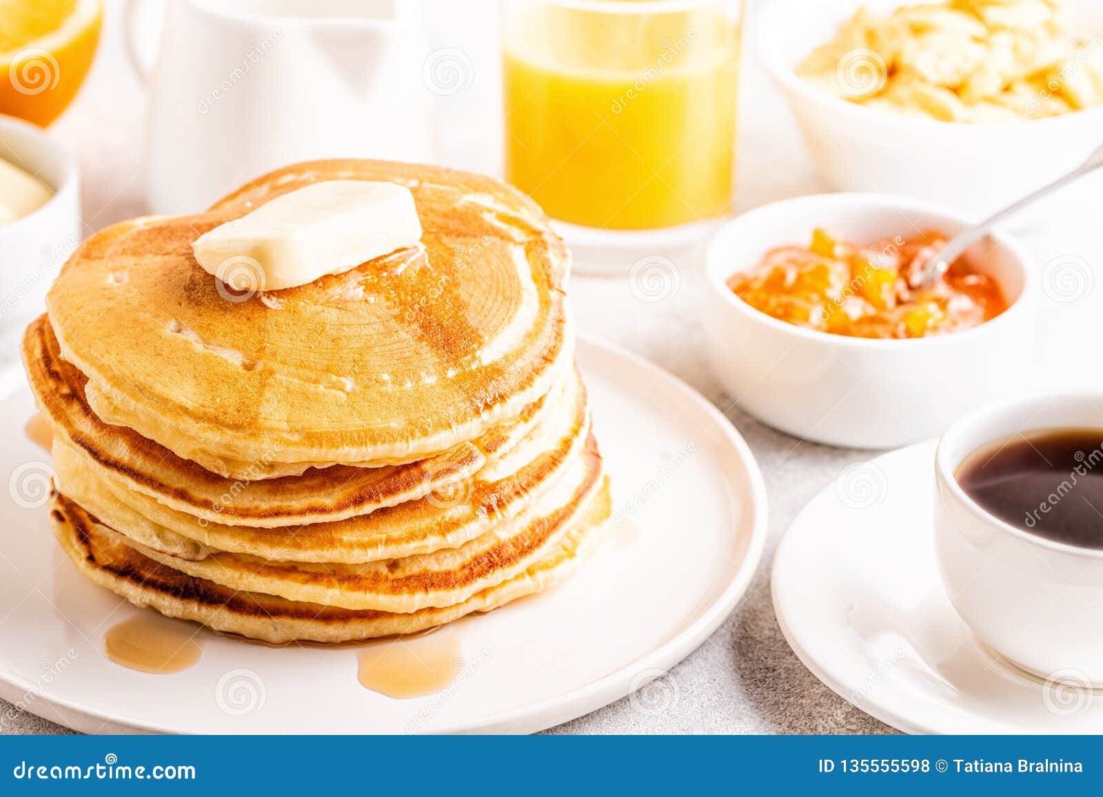 A large stack of fluffy pancakes topped with a pat of soft butter served on a white plate. There are various breakfast items served in white serving dishes nearby. 