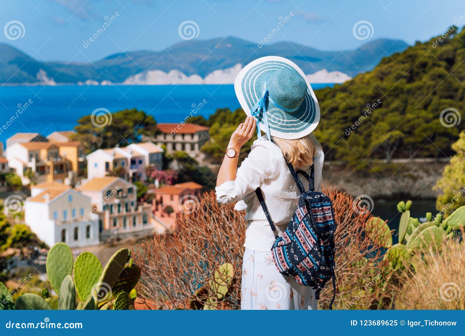 Den turist- kvinnan med blå sunhat, vitkläder och loppryggsäcken beundrar på sikt av den färgrika stillsamma byn Assos. Den turist- kvinnan med blå sunhat, vitkläder och loppryggsäcken beundrar sikt av den färgrika stillsamma byn Assos på solig dag Besöka Kefalonia under sommartid på det Grekland loppet semestra