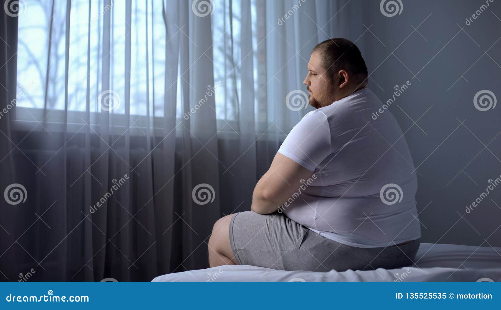 depressed fat man sitting on bed at home, worried about overweight, insecurities