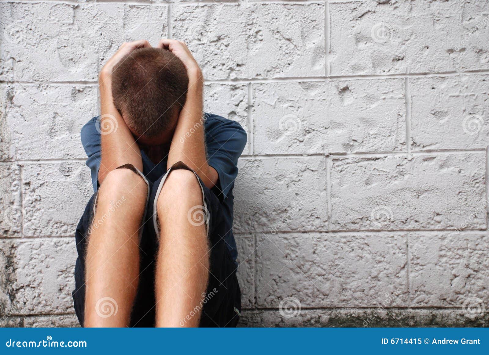 Depressed On The Streets. A young man crouching with his head in his hands, looking down and sitting on a city sidewalk.