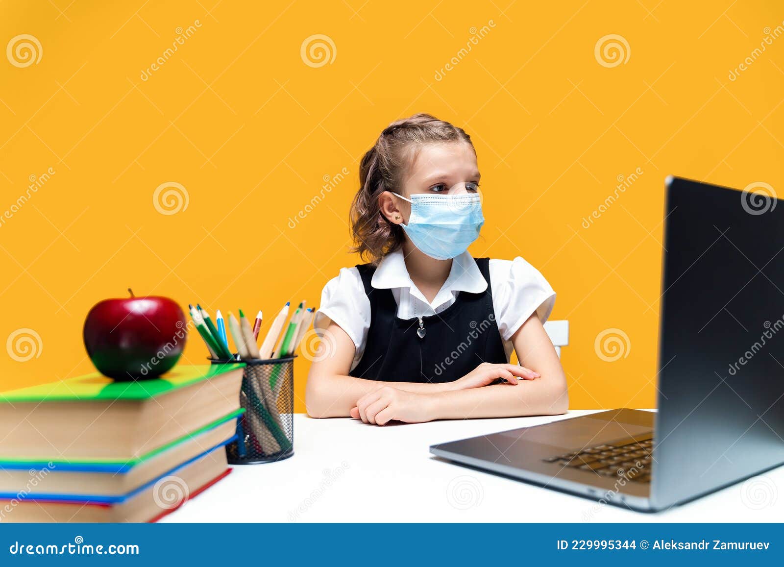 schoolgirl sitting at laptop wearing mask. studying online. yellow background. distance learning.