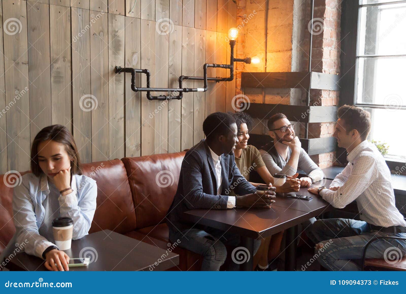diverse young friends ignoring sad girl sitting alone in cafe