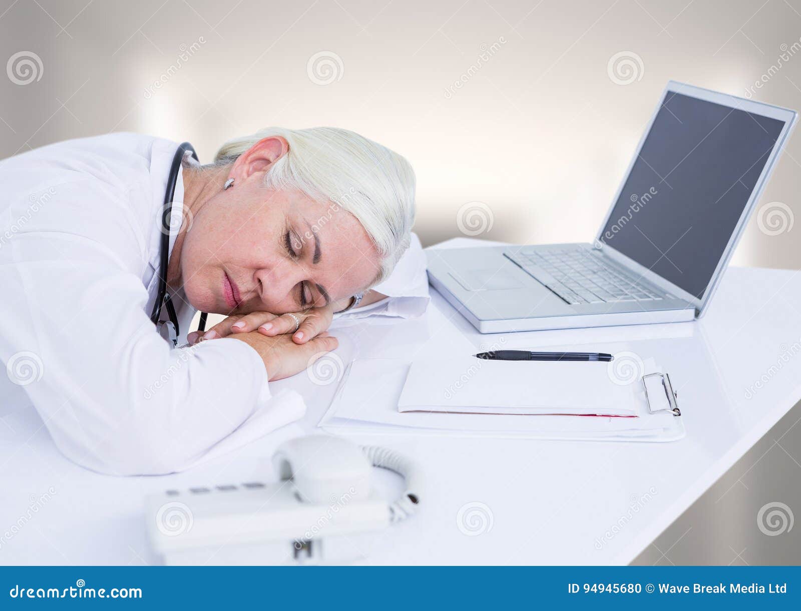 Doctor Asleep At Desk Against White Blurred Hallway Stock Photo