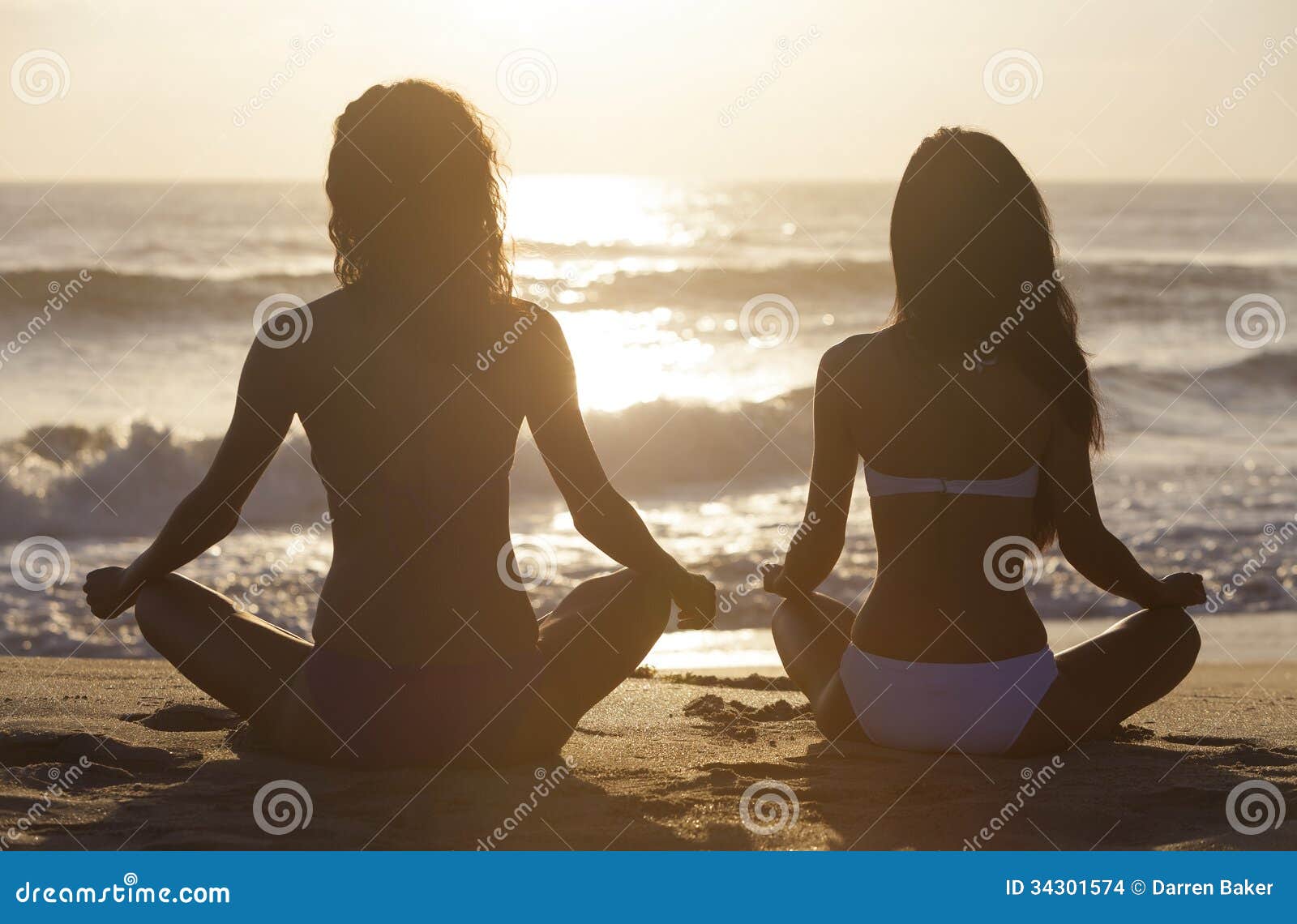 Dos muchachas de las mujeres del bikini que sientan la playa de la salida del sol de la puesta del sol. Dos mujeres jovenes o muchachas atractivas que llevan los bikinis que sientan legged cruzado en una playa tropical abandonada en la puesta del sol o la salida del sol