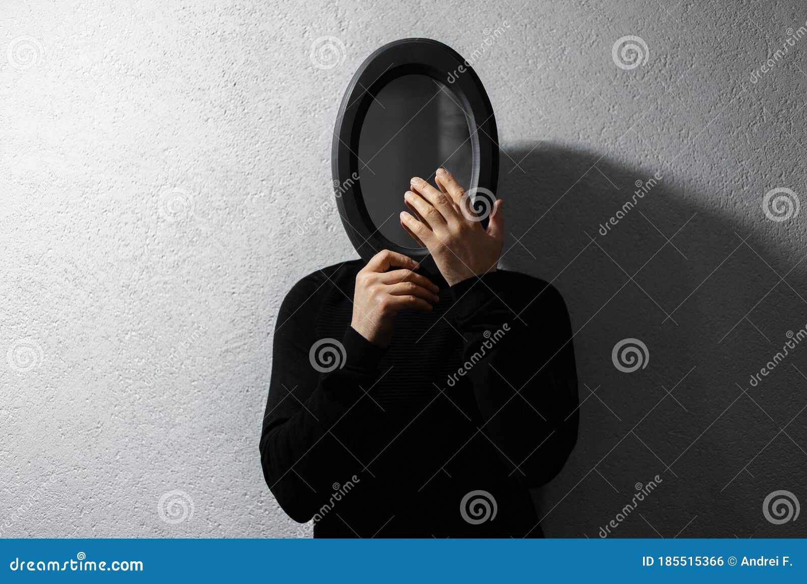 dramatic portrait of young thoughtful guy, looking at his reflection in black oval mirror on textured wall of grey color.