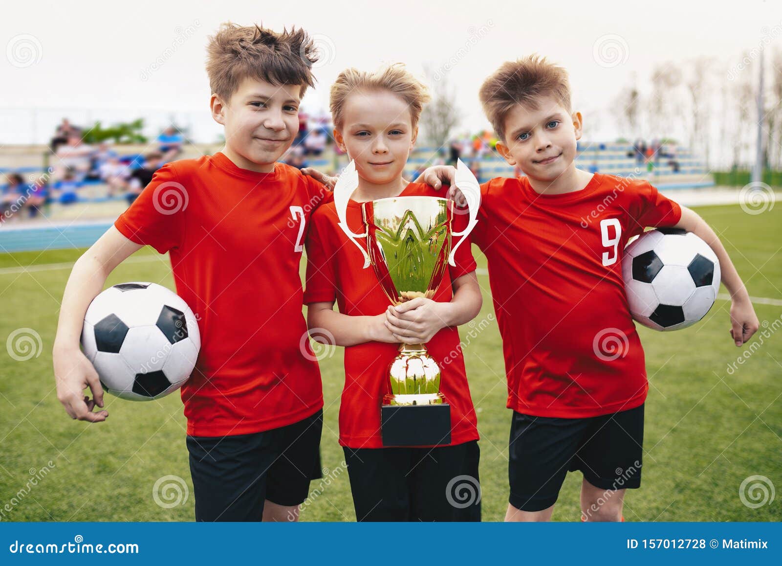 Drei glückliche, fröhliche Kinder des Sportfußballteams Boys Football Players Holding Trophy im Stadion Junge Gewinner des Jugendfußballturniers