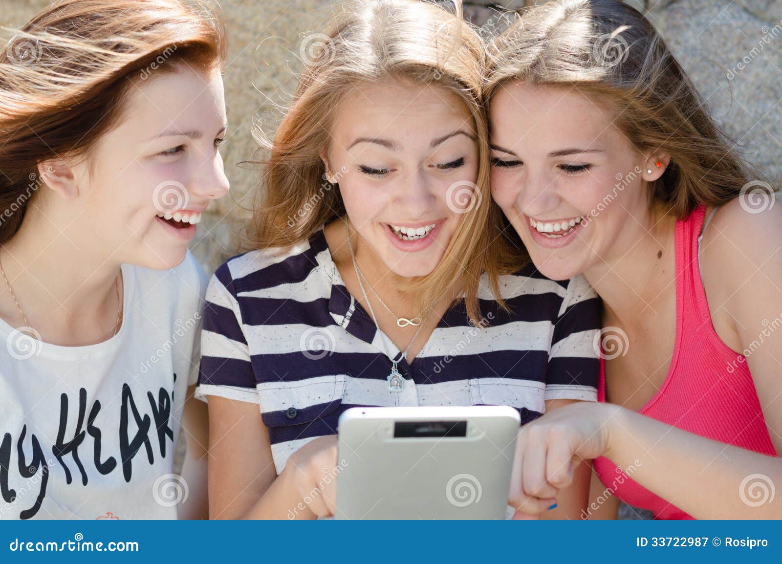 Drie gelukkige vrienden van het tienermeisje en tabletcomputer. Drie gelukkige vrienden die van het tienermeisje op tabletpc kijken terwijl het zitten op groen gazon op de zomerdag