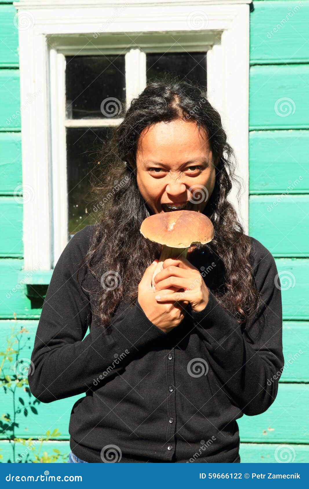 Dziewczyny mienia pieczarka. Indonezyjska dziewczyna - uśmiechnięta młoda kobieta gryźć dużej pieczarki typ borowik w czarnym ciągnieniu (grzyb)