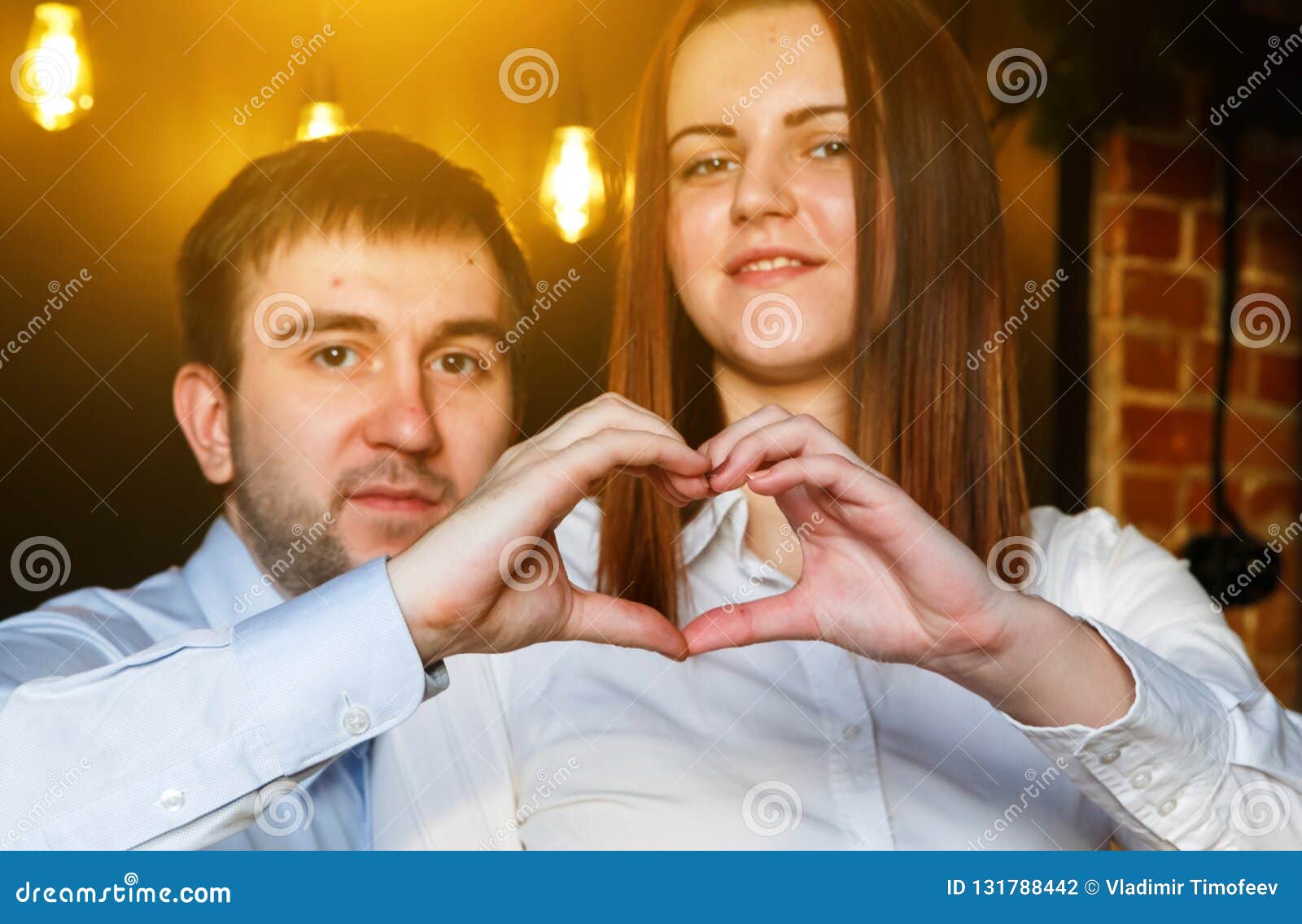Happy couple in love showing heart with fingers in the loft interior, burning lights in the background