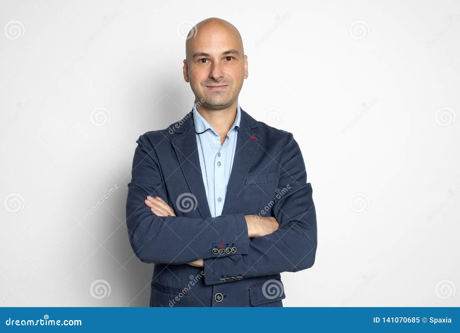 Cheerful middle aged bald handsome man keeping arms crossed and looking at camera with smile while standing grey background