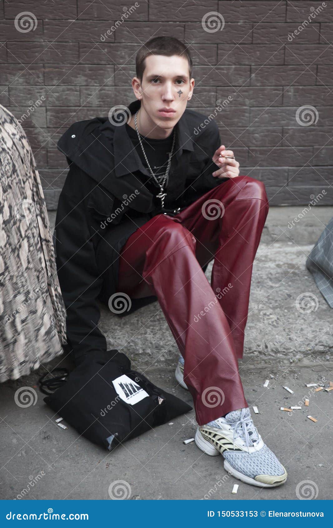 Moscow, RUSSIA - June 12, 2019: A young man in a black jacket, burgundy leather pants, and a cross-shaped tattoo on his face is sitting on the sidewalk