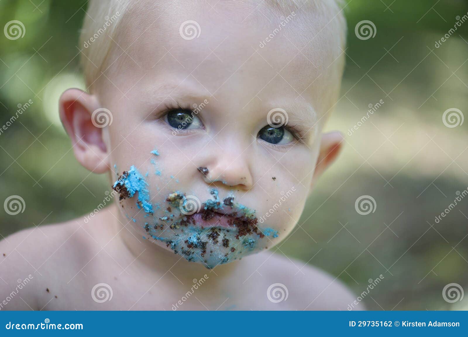 Kuchenzertrümmerntrieb: Baby, das Kuchen auf seinen Tannen isst. Ein Baby mit einem unordentlichen Gesicht genießt einen blauen und orange Kuchen draußen zur Feier seines ersten Geburtstages