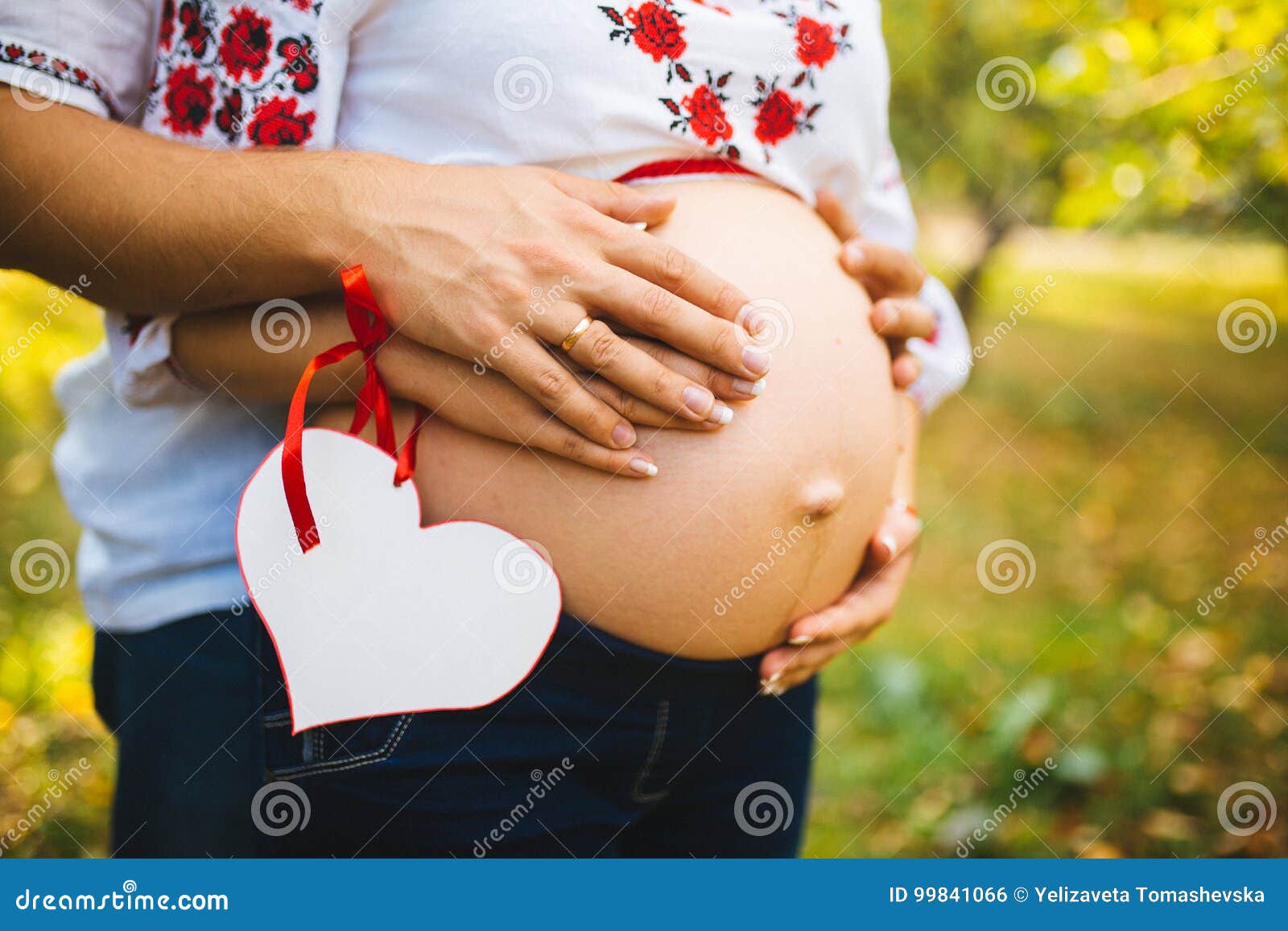 Eine schwangere Frau mit ihrer Ehemannumarmung der Bauch, gekleidet in einem traditionellen ukrainischen Ausstattungs- und Griffp. Eine schwangere Frau mit ihrer Ehemannumarmung der Bauch, gekleidet in einer traditionellen ukrainischen Ausstattung und halten ein Papierherz mit Raum für Text