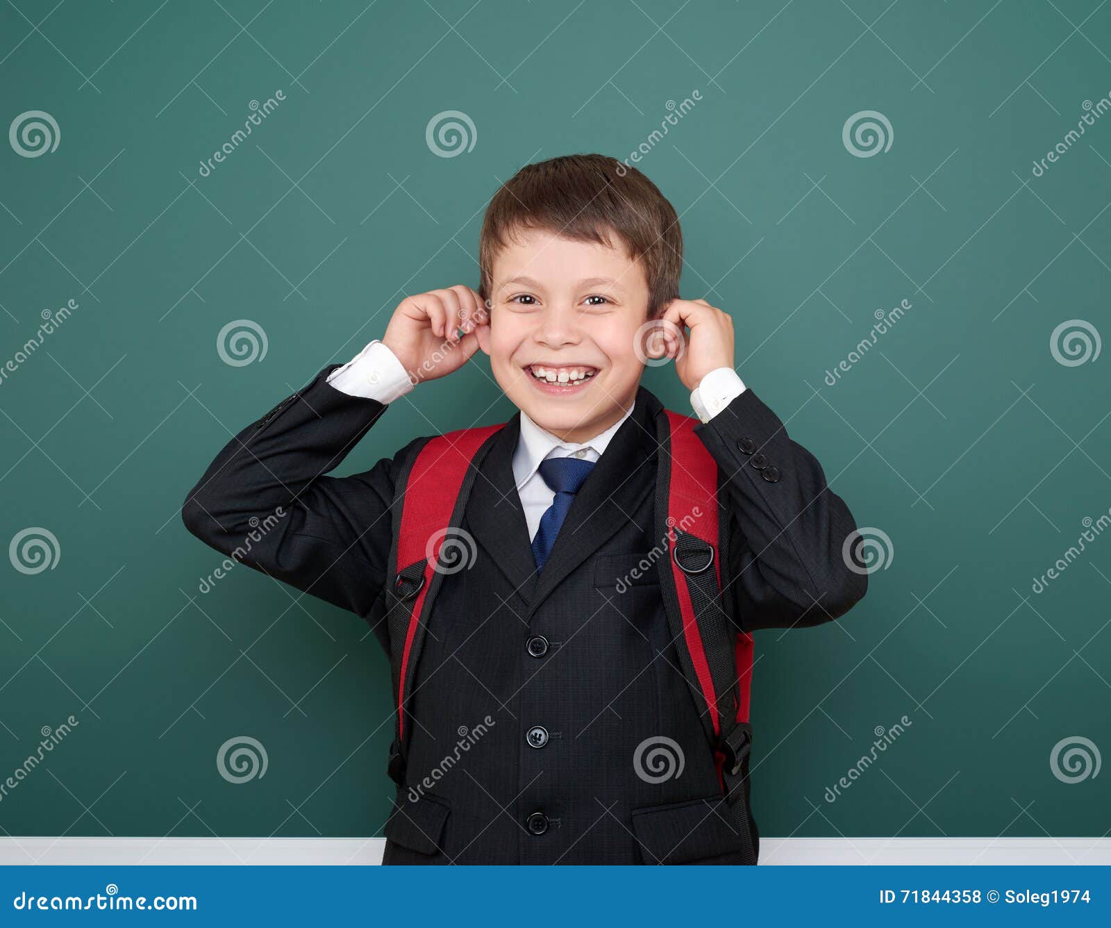 El escolar hace caras en traje negro en el fondo verde con la mochila roja, concepto de la pizarra de la educación