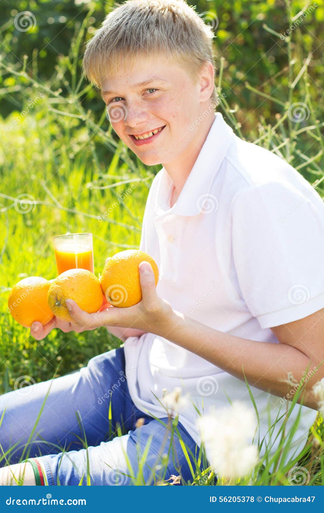 El muchacho del adolescente se está sosteniendo de cristal con el zumo de naranja. El retrato del muchacho sonriente rubio del adolescente con las pecas en su cara se está sosteniendo de cristal con el zumo de naranja y las frutas, tiempo de verano