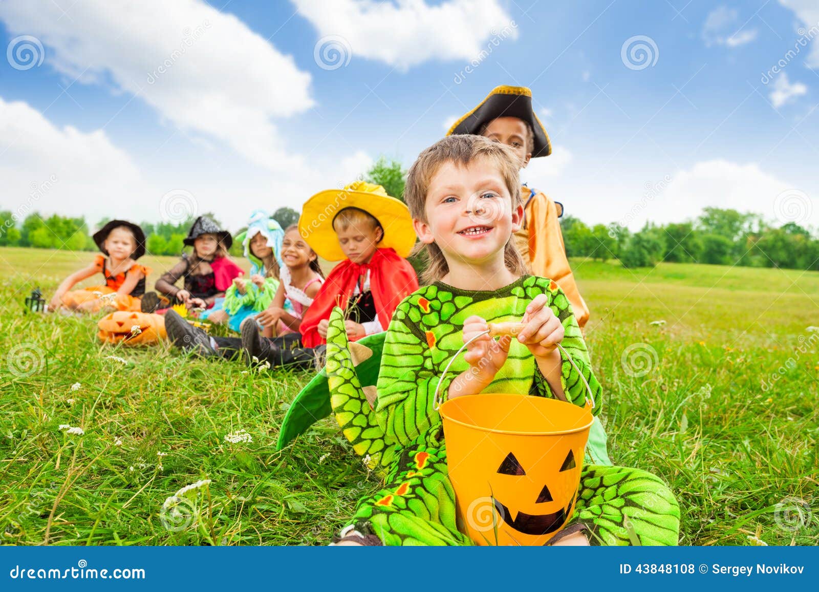 El muchacho lindo en traje del monstruo sostiene el cubo de Halloween. El muchacho sonriente lindo en traje del monstruo sostiene el cubo de Halloween y sonríe con sus amigos que se sientan en fondo en fila