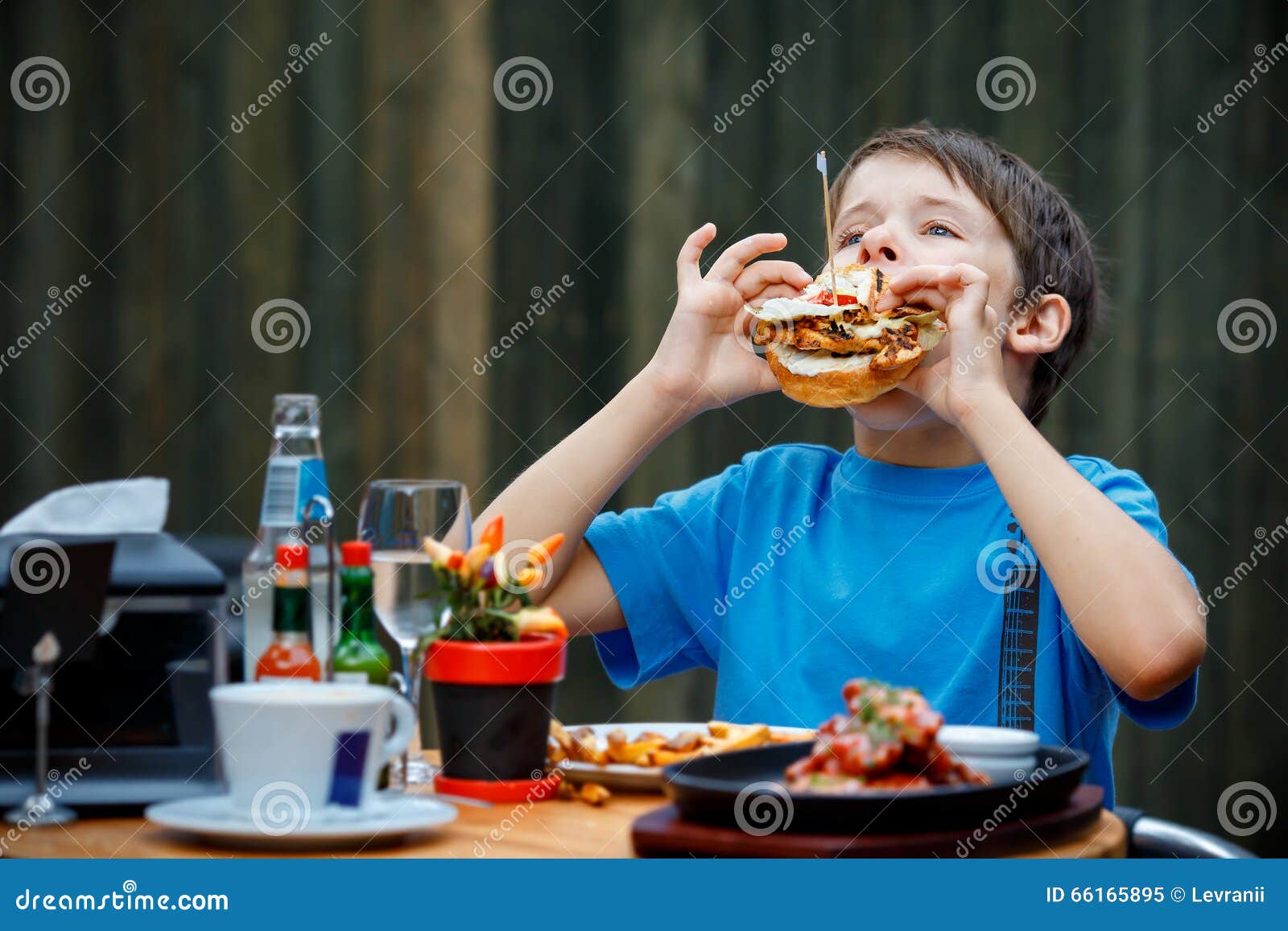 El muchacho sano lindo del adolescente come la hamburguesa y la patata que se sientan en café al aire libre