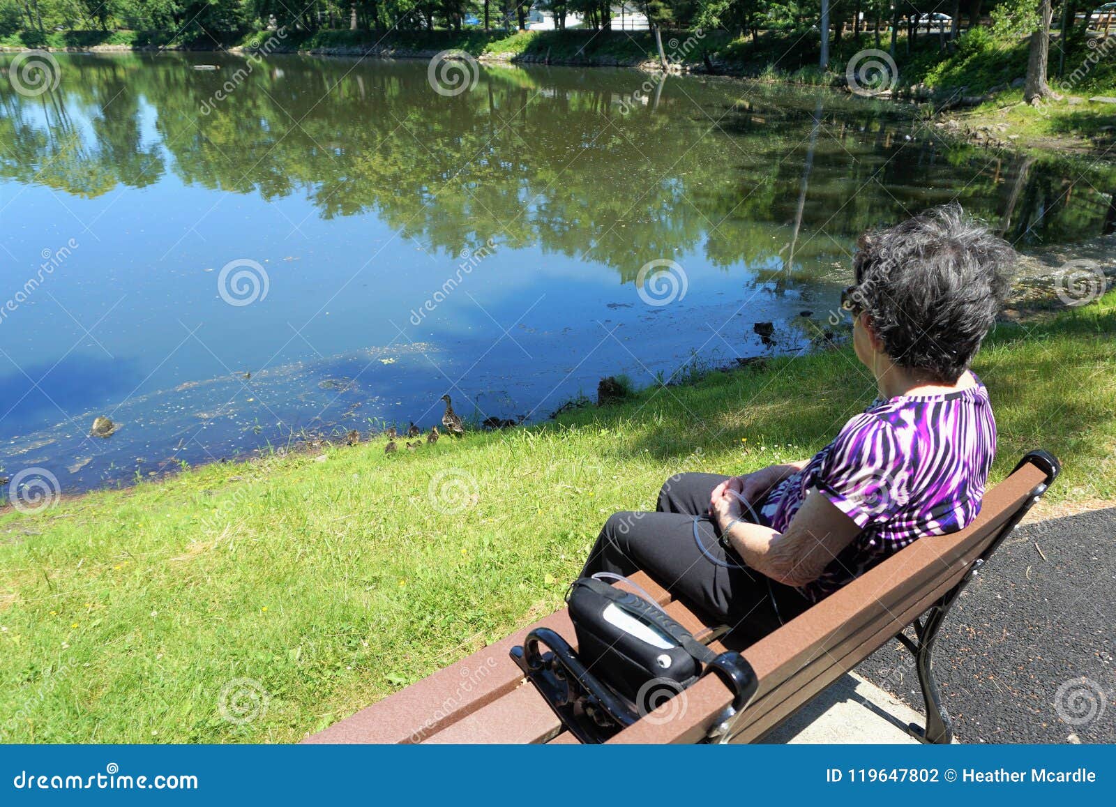 older woman sits on bench with oxygen tank looking at pond