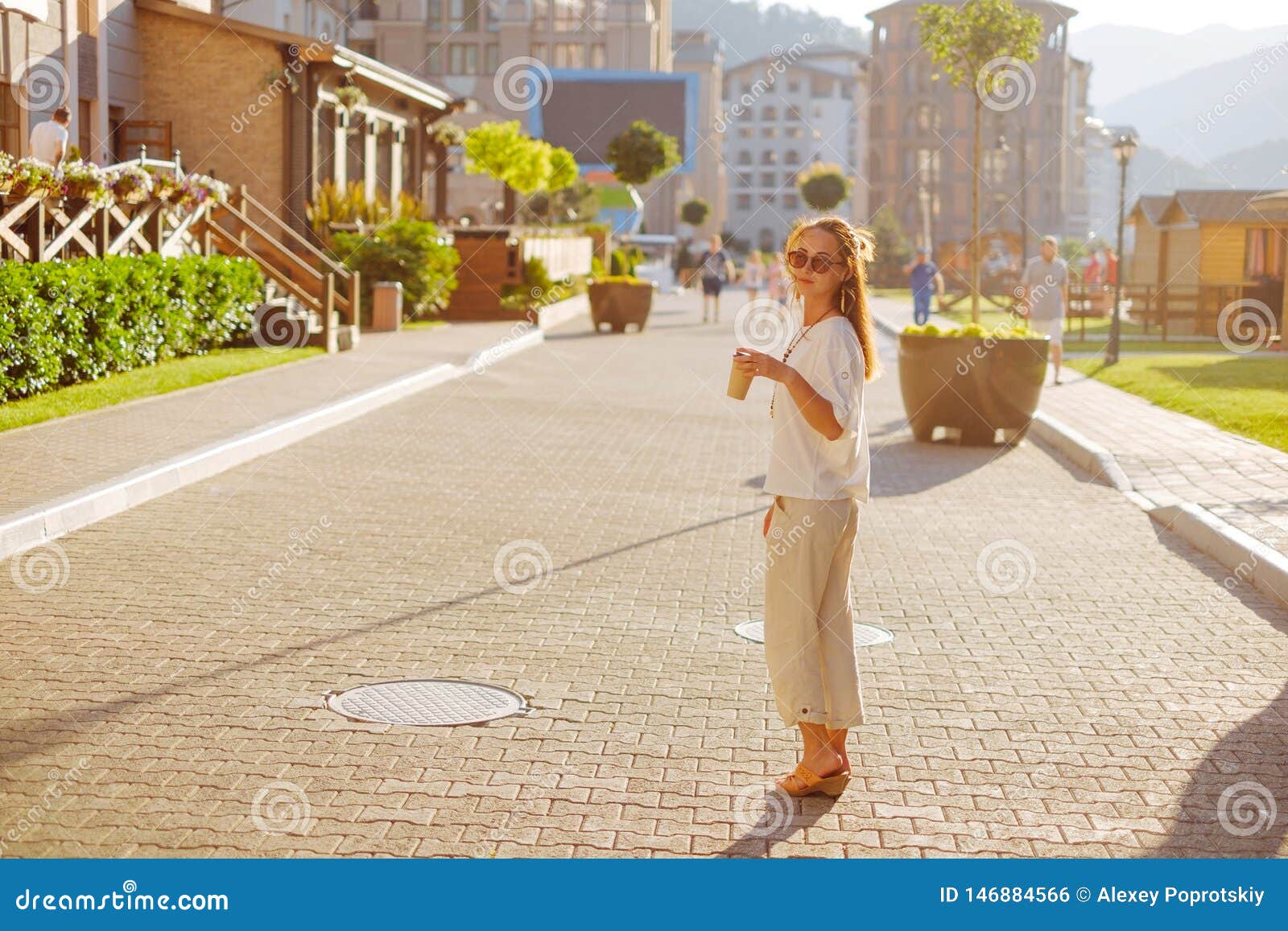 Elegancki kobiety odprowadzenie z filiżanka kawy na ulicie. Elegancka młoda kobieta jest ubranym w białym ubraniowym odprowadzeniu na ulicie z papierowym filiżanka kawy w ranku, miasta styl życia