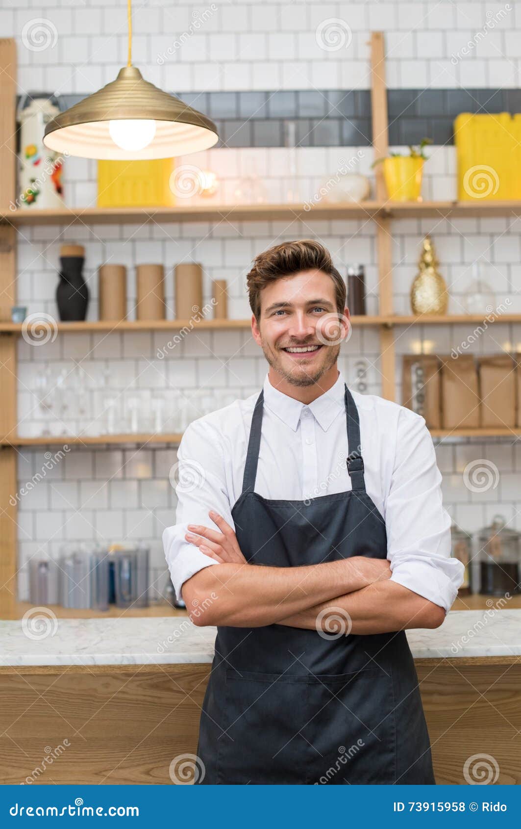 Empresario joven feliz. Retrato de un camarero joven sonriente que se inclina en el contador de la cafetería con los brazos cruzados Dueño acertado con el delantal que mira la cámara Cocinero de los jóvenes que se coloca en cocina concepto de la pequeña empresa