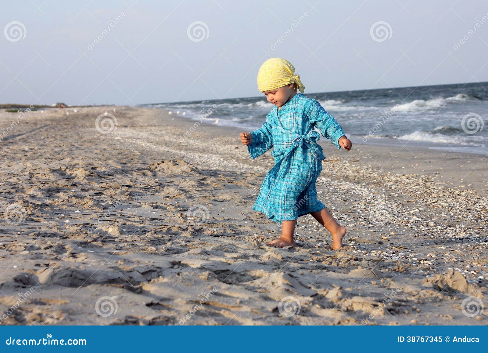 Enfant en bas âge sur la plage. L'enfant en bas âge marchant sur la plage s'est habillé avec une robe bleue et un bandana jaune, recherchant des coquillages.