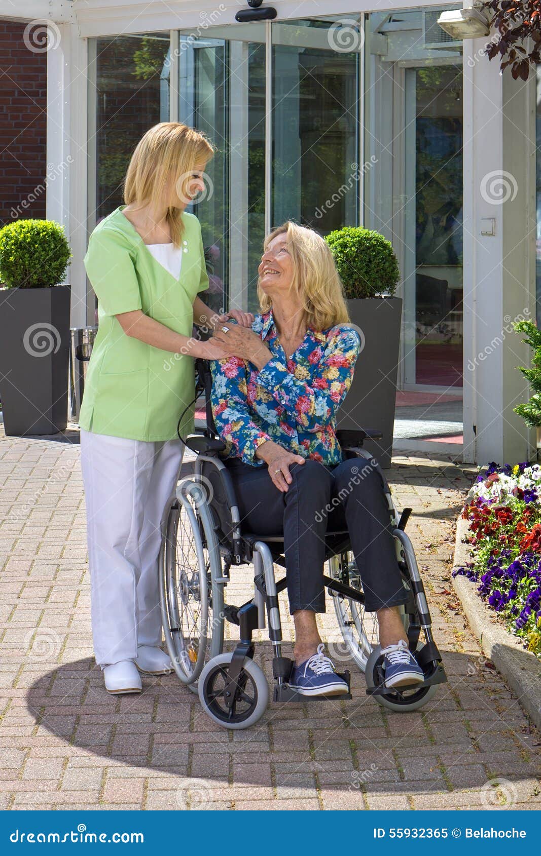 Enfermera Showing Care para la mujer mayor en silla de ruedas. Enfermera rubia que cuida Standing con la mano en el hombro de la mujer mayor que se sienta en silla de ruedas al aire libre delante del edificio en Sunny Day