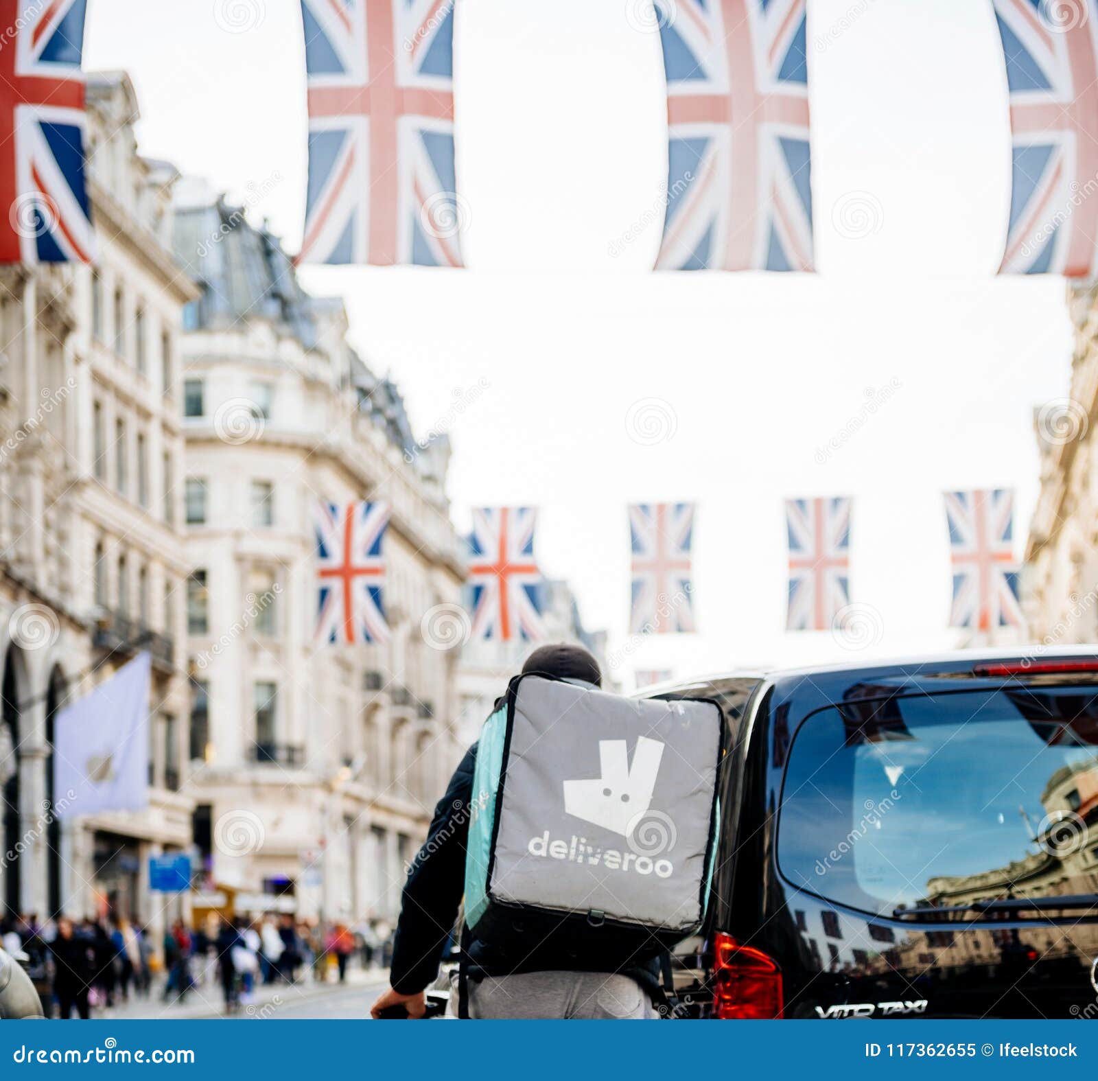 Entrega do alimento de Deliverro em Londres durante o casamento real Regent St. LONDRES, REINO UNIDO - 18 DE MAIO DE 2018: Imagem quadrada do ciclista de Deliveroo que espera na luz vermelha perto do táxi em Regent Street com bandeiras que comemora a união de Meghan Markle Prince Harry