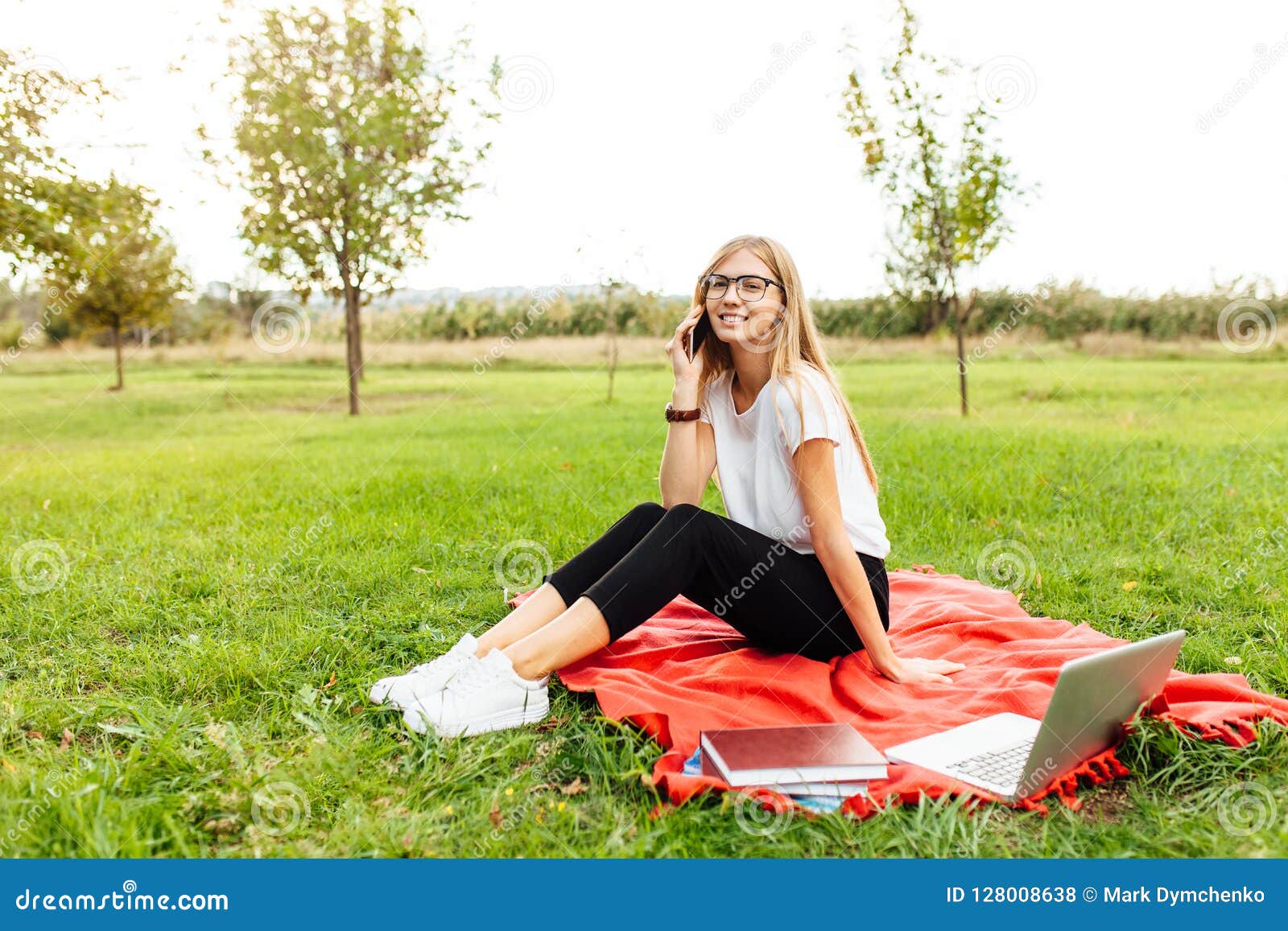 Estudante bonito novo com os vidros, falando no telefone, e. Imagem de um estudante bonito novo com vidros, falando no telefone, e sentando-se em uma cobertura vermelha no parque, com livros e um portátil