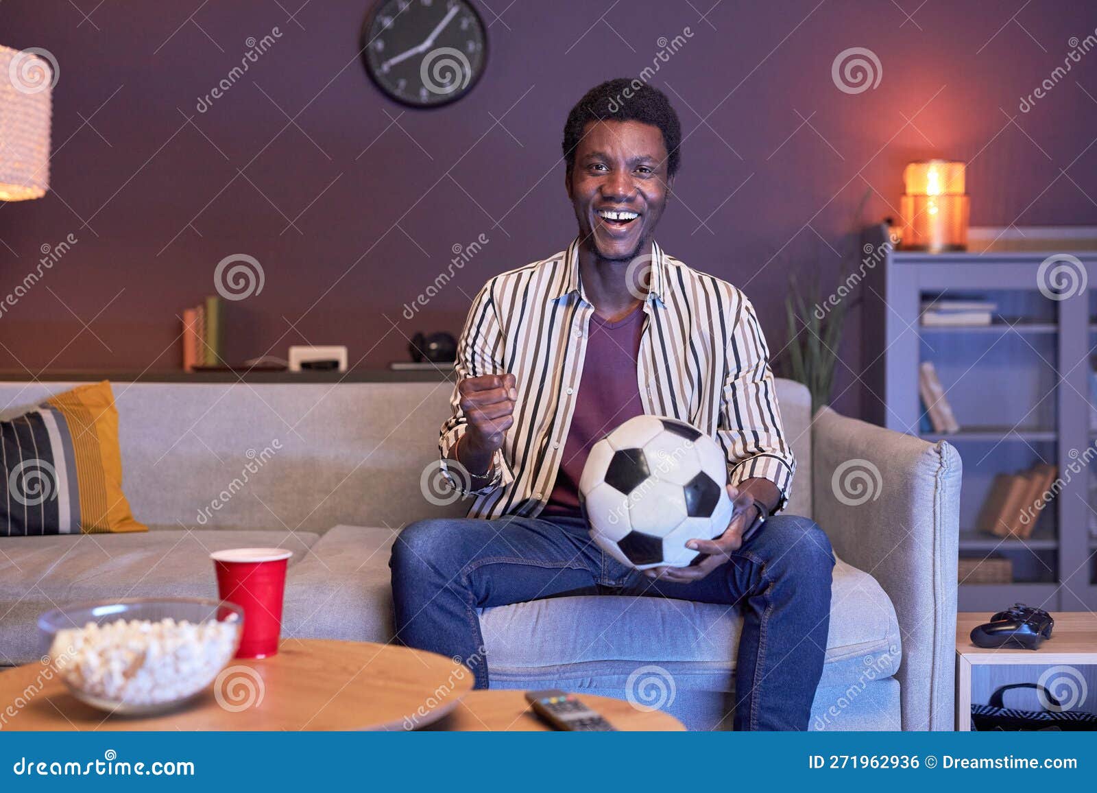 Excited Black Man Watching Football Match on Tv at Home and Holding Ball Stock Photo