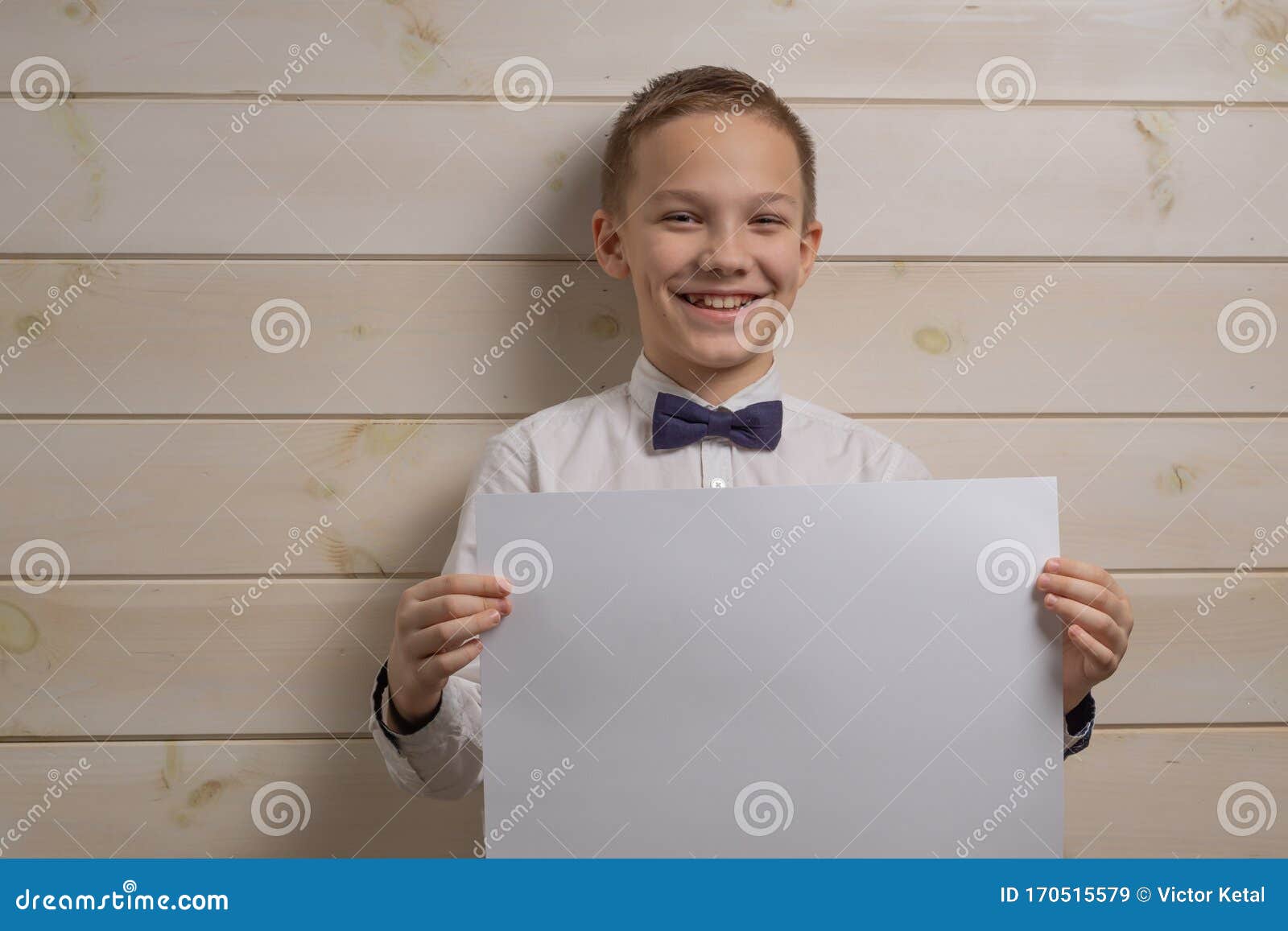 a fair-haired boy of 10 years in a white shirt with the self-tie bow tie smiles against the background of a wooden wall. the boy