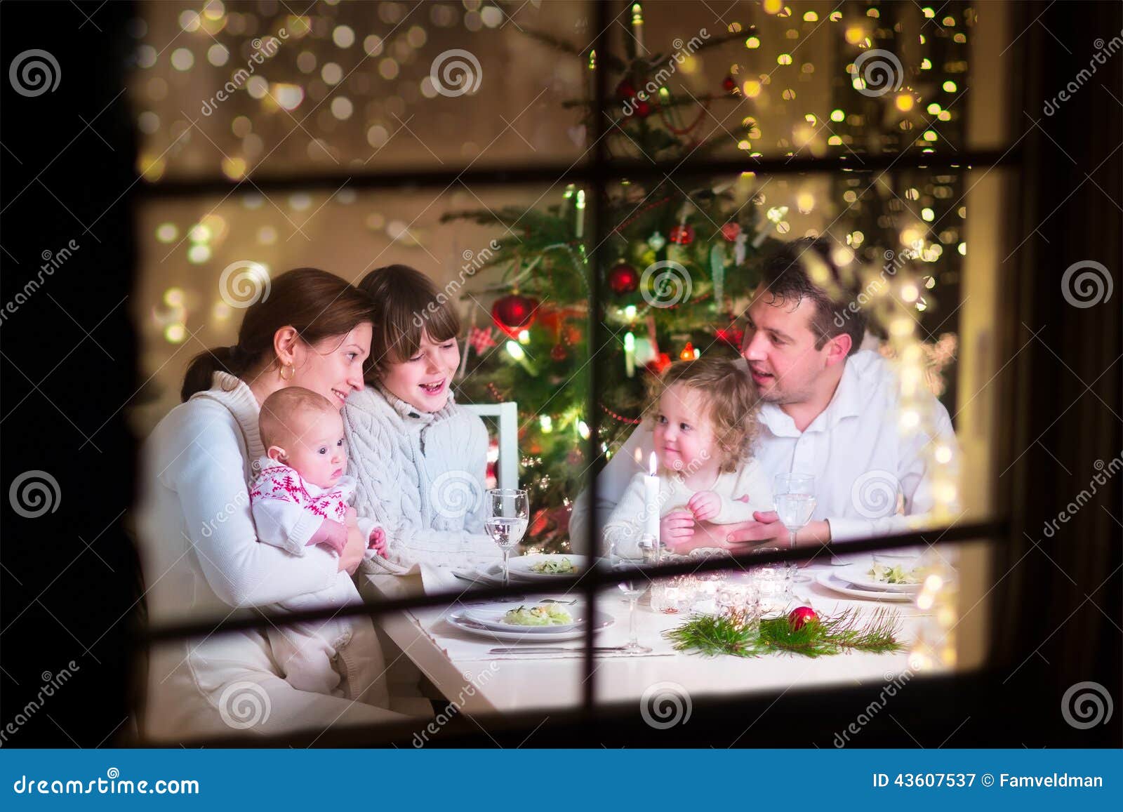 Familia en la cena de la Navidad. La familia grande joven que celebra la Navidad que disfruta de la cena, la visión desde el exterior a través de una ventana en una sala de estar adornada con el árbol y la vela se enciende, los padres felices que comen con tres niños