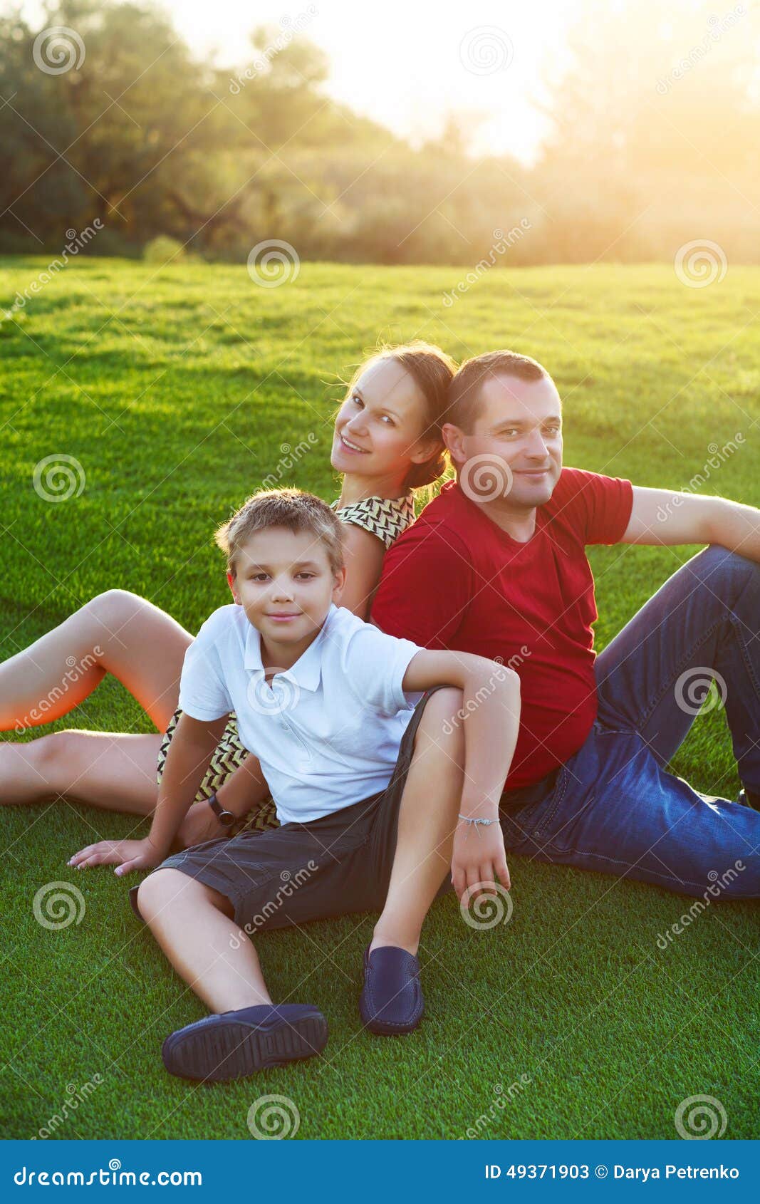 Familia feliz con el hijo que se sienta en hierba en el parque Puesta del sol del verano