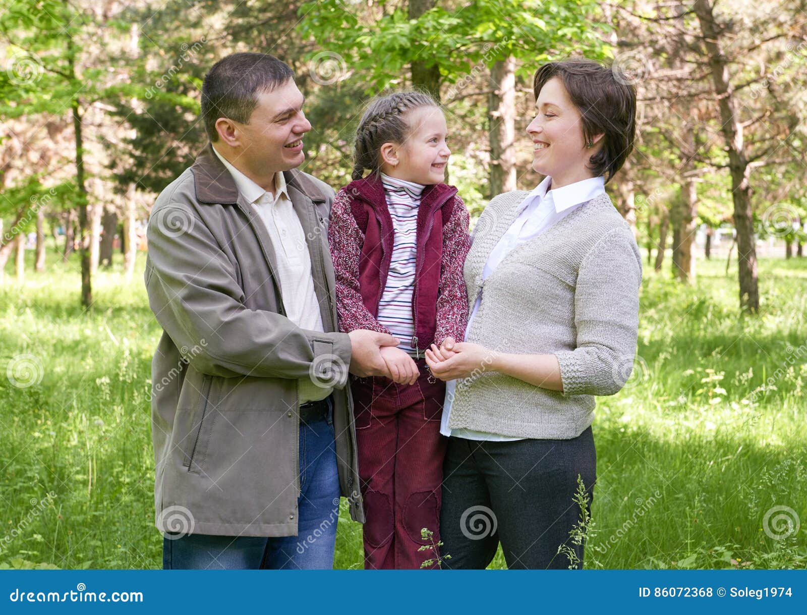 Familia feliz con el niño en parque del verano, luz del sol, hierba verde y árboles