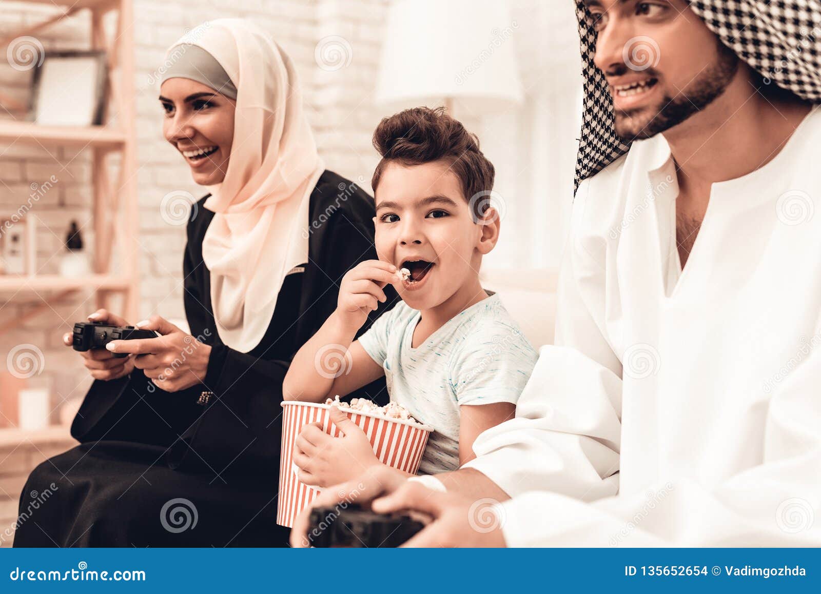 Familia árabe feliz que juega en la consola en casa Familia que se sienta en el sofá Hombre que usa el dispositivo de Digitaces Boay sonriente en casa E Muchacho sonriente Mujer árabe joven Mujer en vestido negro