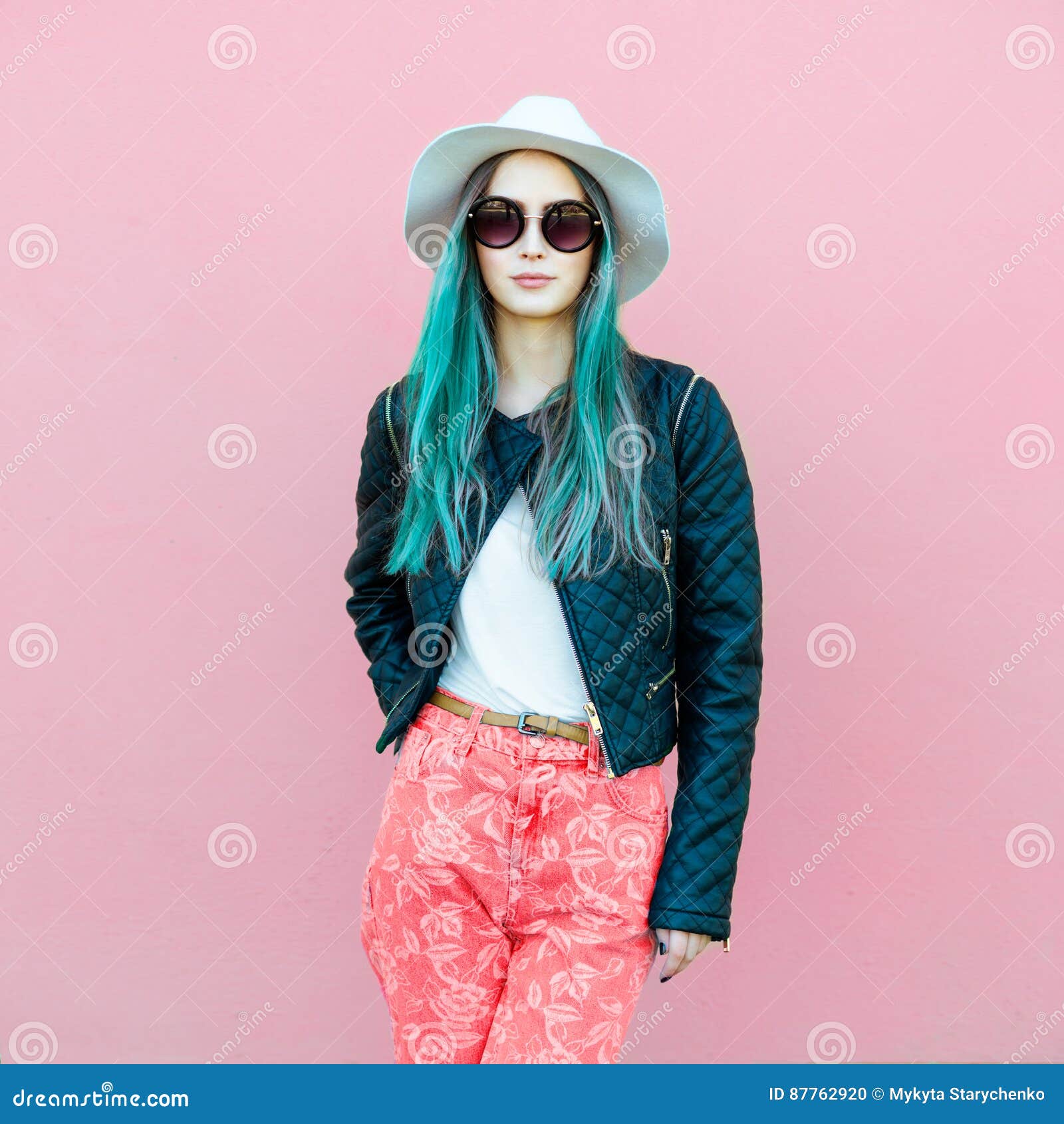 fashionable young blogger woman with blue hair wearing casual style outfit with black jacket, white hat, pink jeans and sunglasses