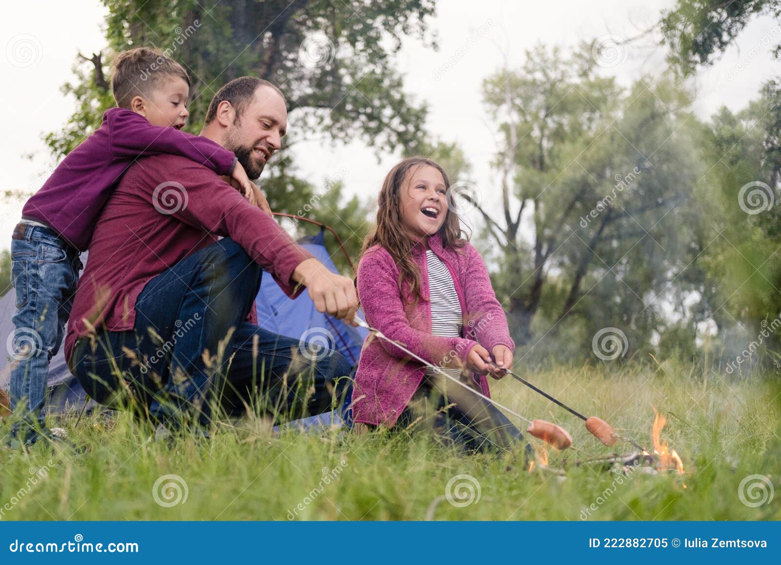 father, daugther and son roasting sausages at campfire in forest