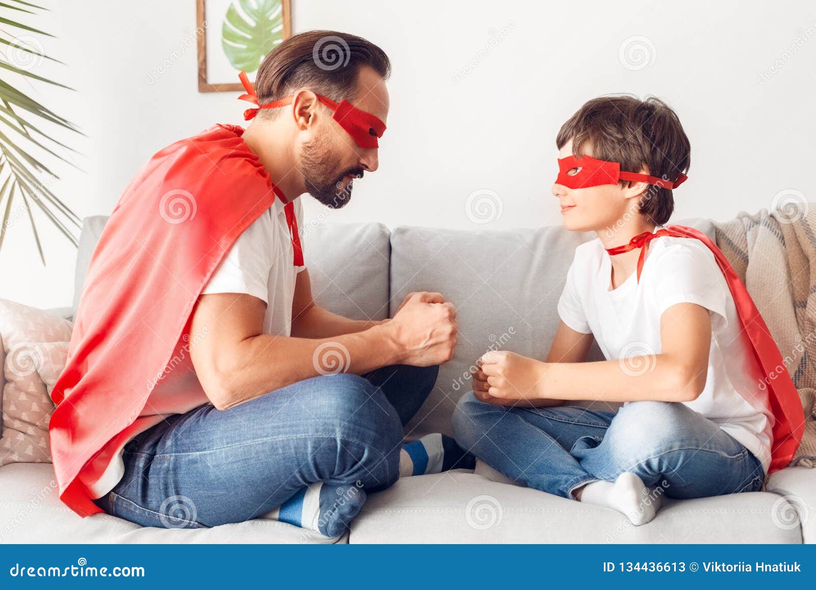 father and son in superheroe costumes at home sitting on sofa clencing fists playing looking at each other joyful side