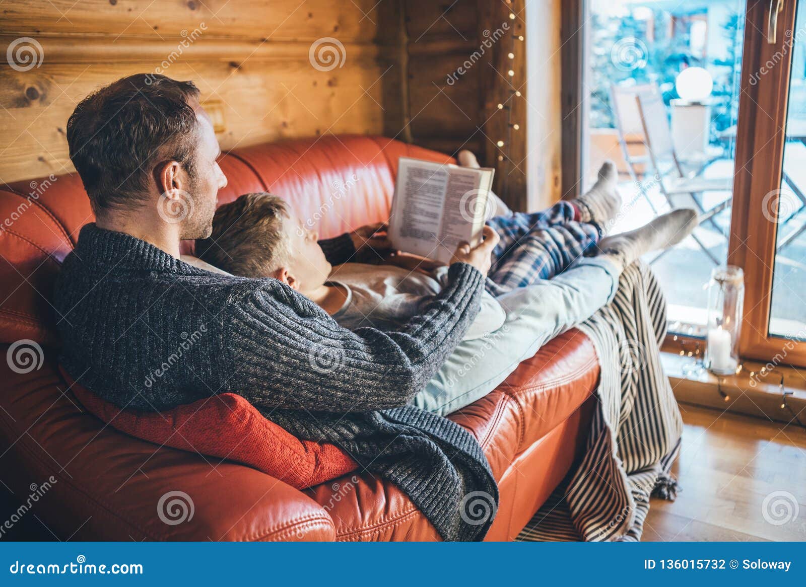 father and son reading book together lying on the cozy sofa in warm country house. reading to kids conceptual image