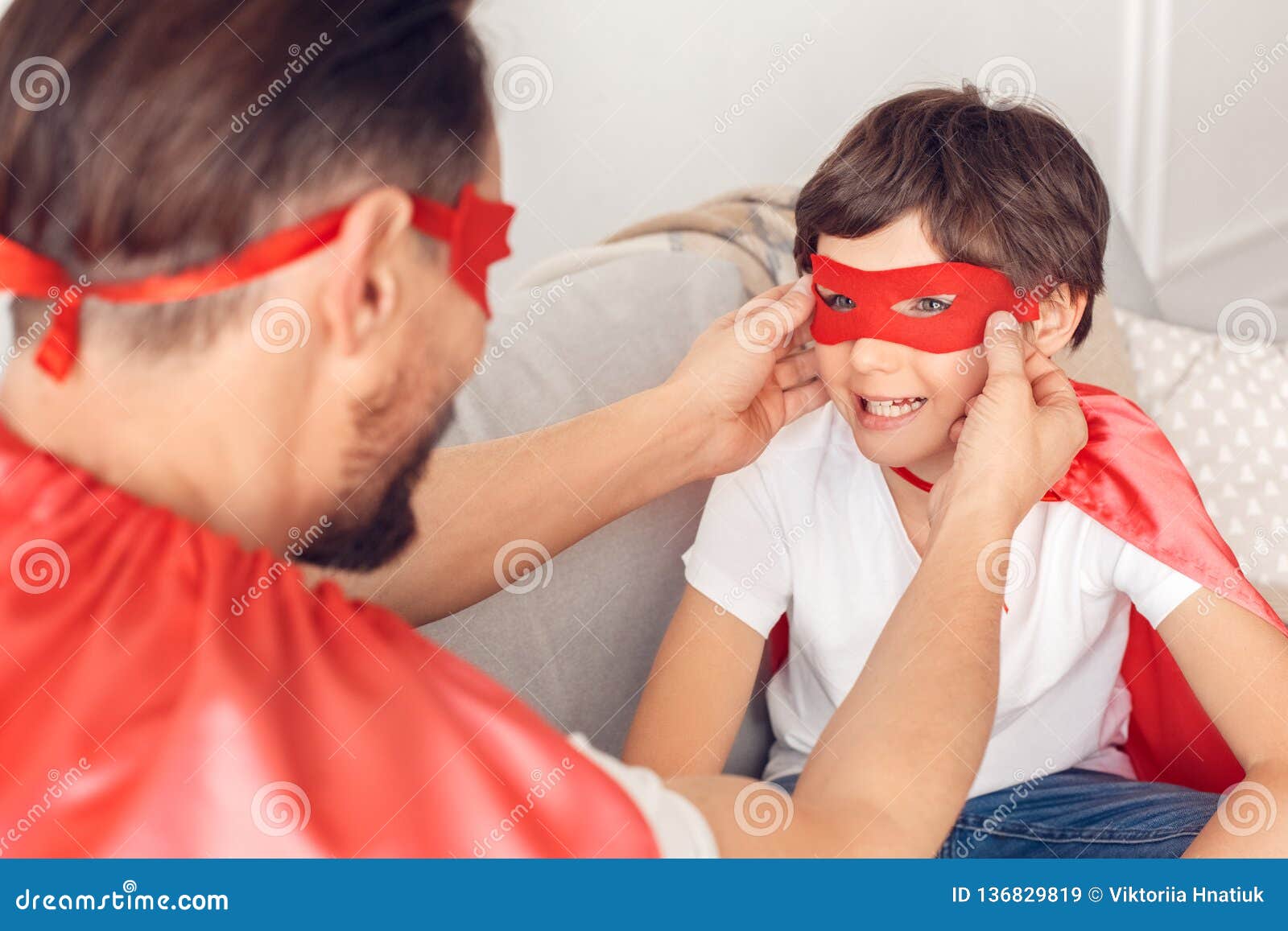 father and son in superheroe costumes at home sitting on sofa man putting mask on boy smiling close-up
