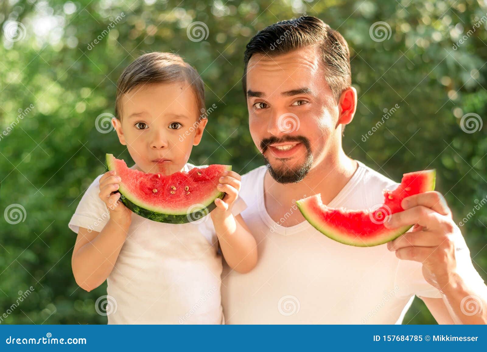 father and son together eating watermelons, both man and kid are holding slices of juicy watermelons, green leaves in background