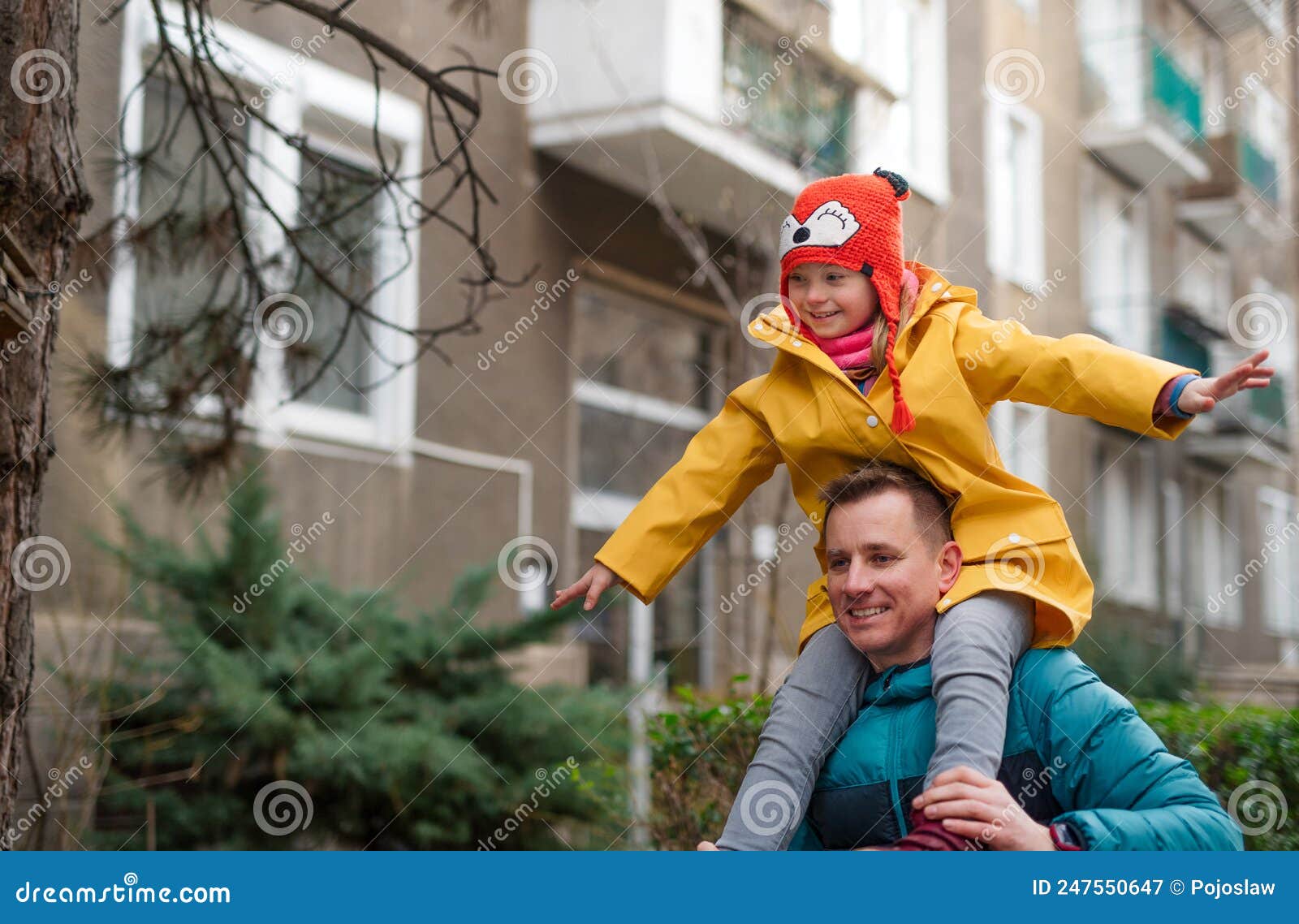 father taking his little daughter with down syndrome on piggyback to school, outdoors in street.