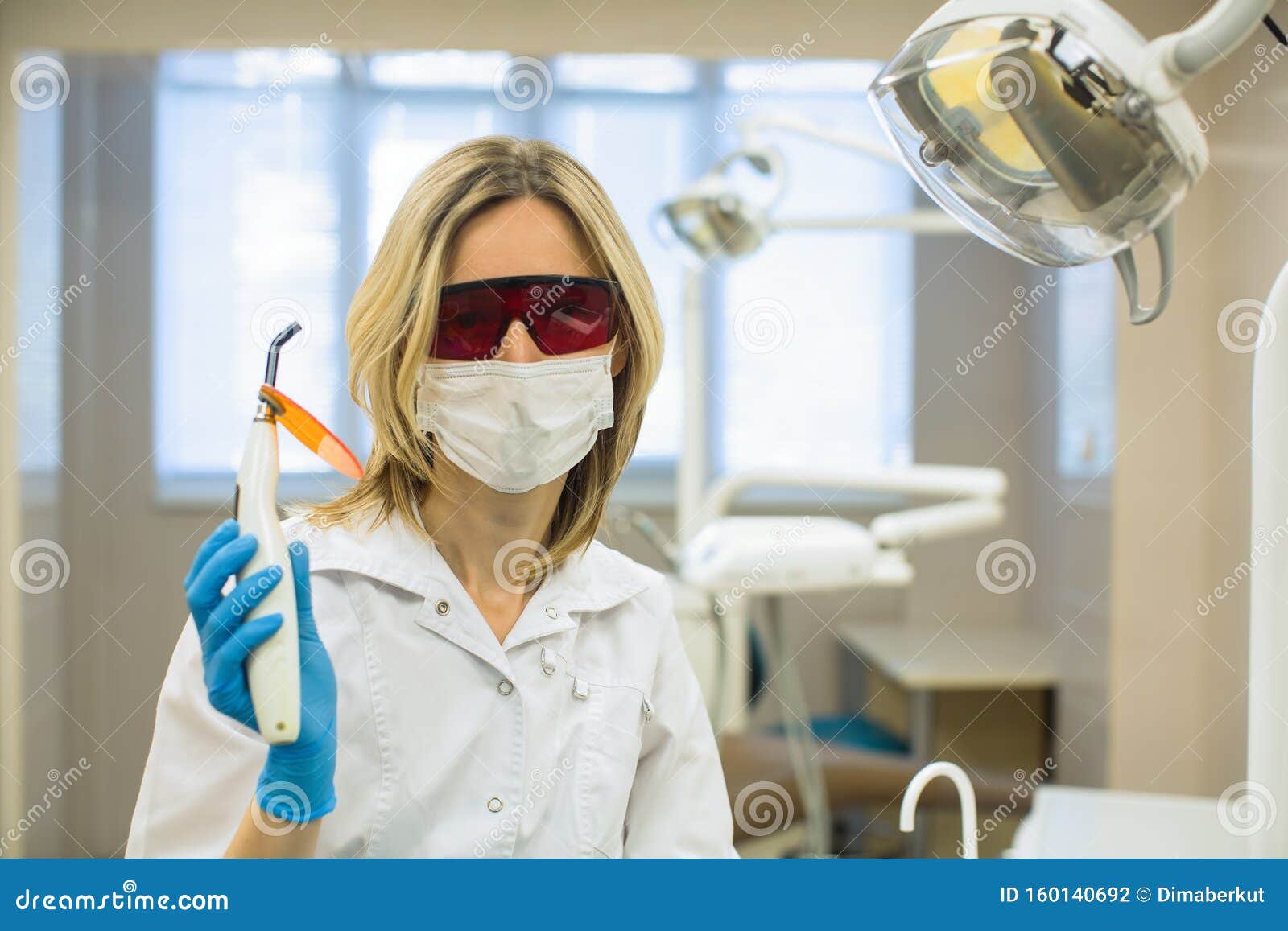 Female Dentist Doctor With Safety Glasses Shows A Dental Curing Light In Dental Office Stock