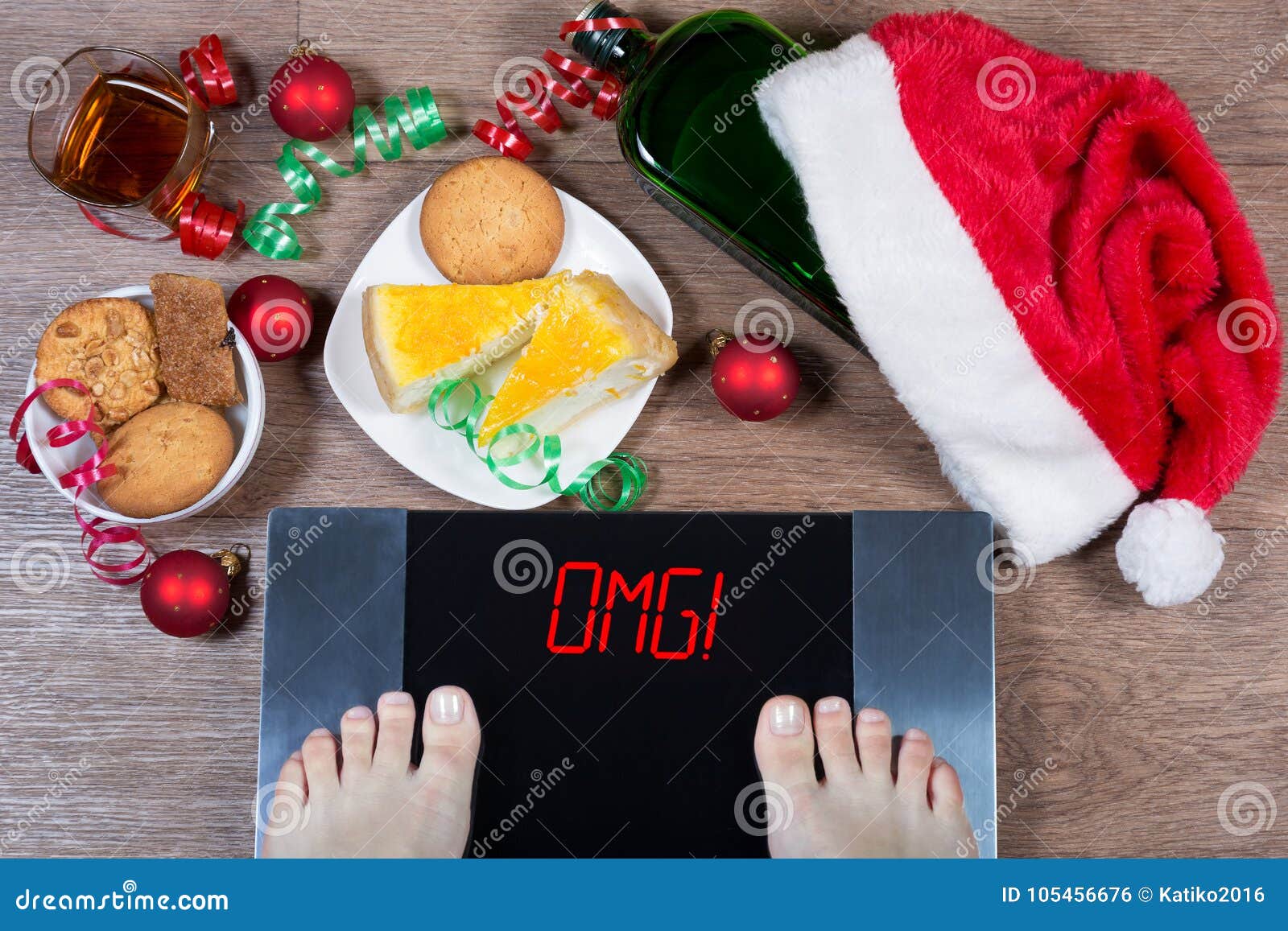 female feet on digital scales with sign omg! surrounded by christmas decorations, bottle, glass of alcohol and sweets.