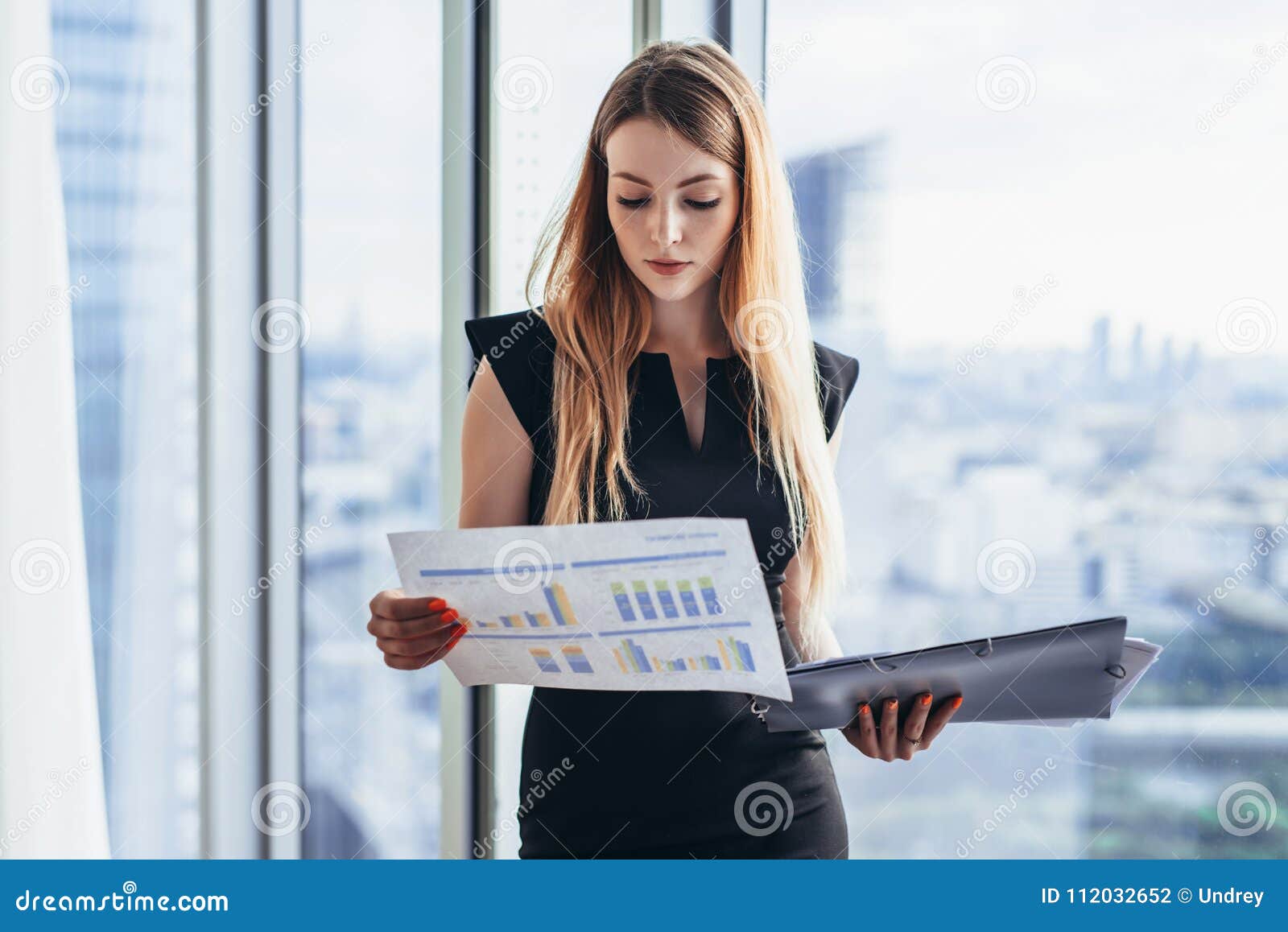 female financial analyst holding papers studying documents standing against window with city view
