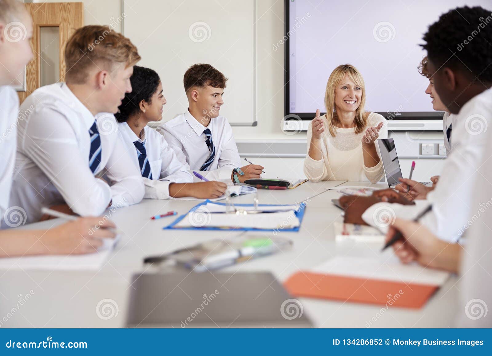 female high school teacher sitting at table with teenage pupils wearing uniform teaching lesson